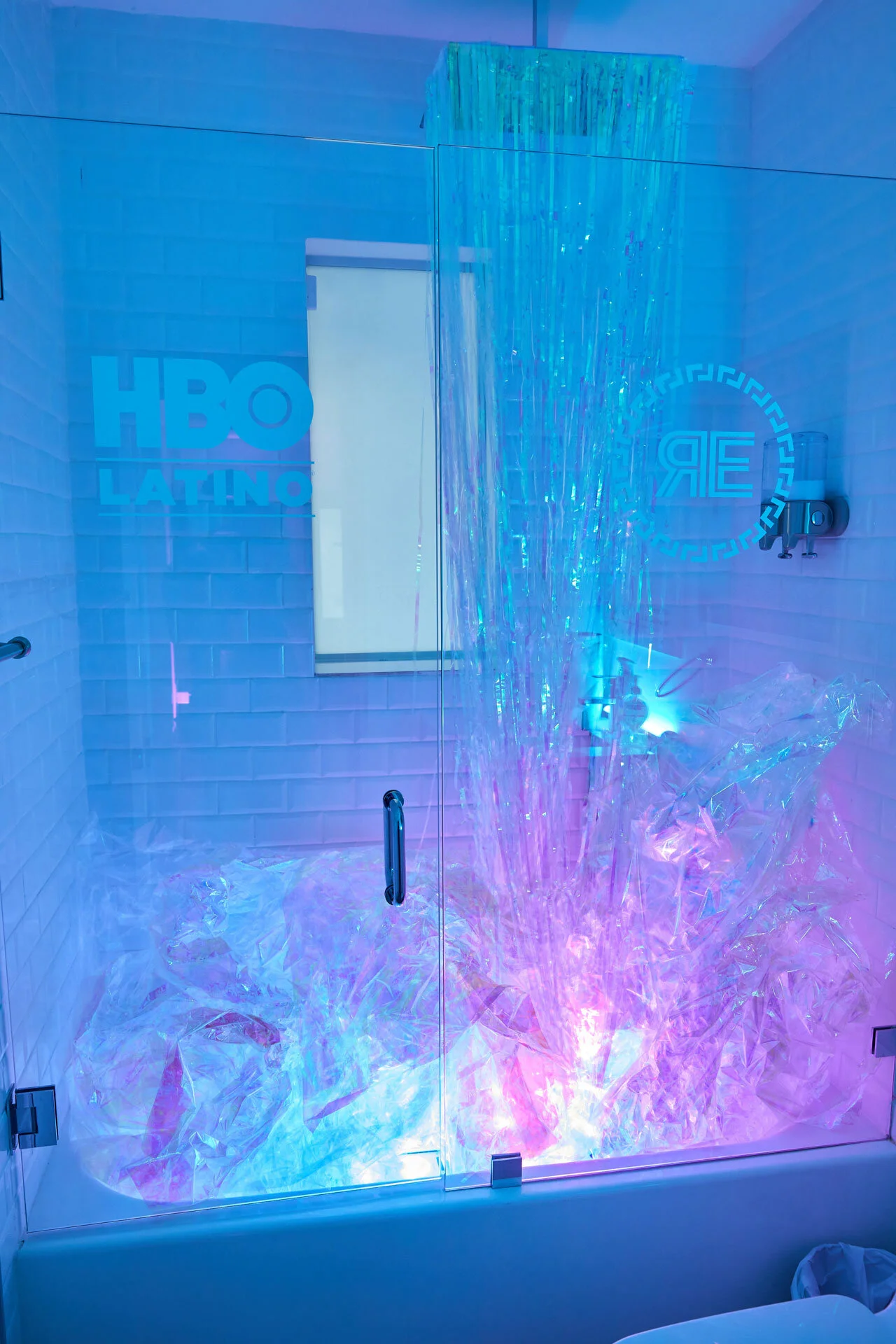 Una ducha con pared de azulejos blancos y una pantalla que dice HBO Latino atrás. La ducha tiene efectos de luces de colores y decoraciones de plástico transparente en el suelo.
