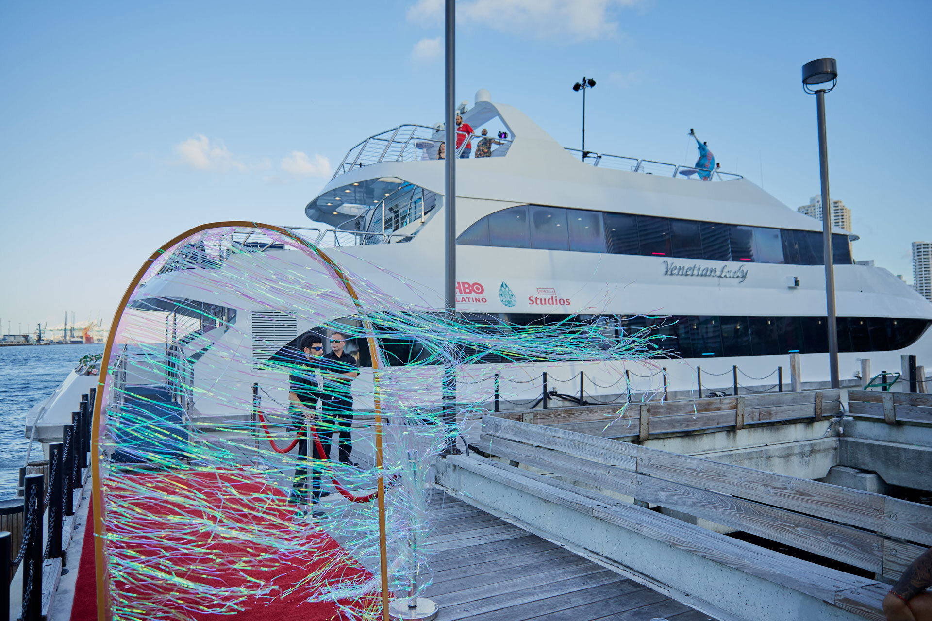 Un yate grande y moderno en el muelle con un arco de bienvenida decorado con luces y una pista roja. El yate tiene logotipos de HBO Latino y Estudios. El mar y el cielo azul están en el fondo, con edificios en la distancia.