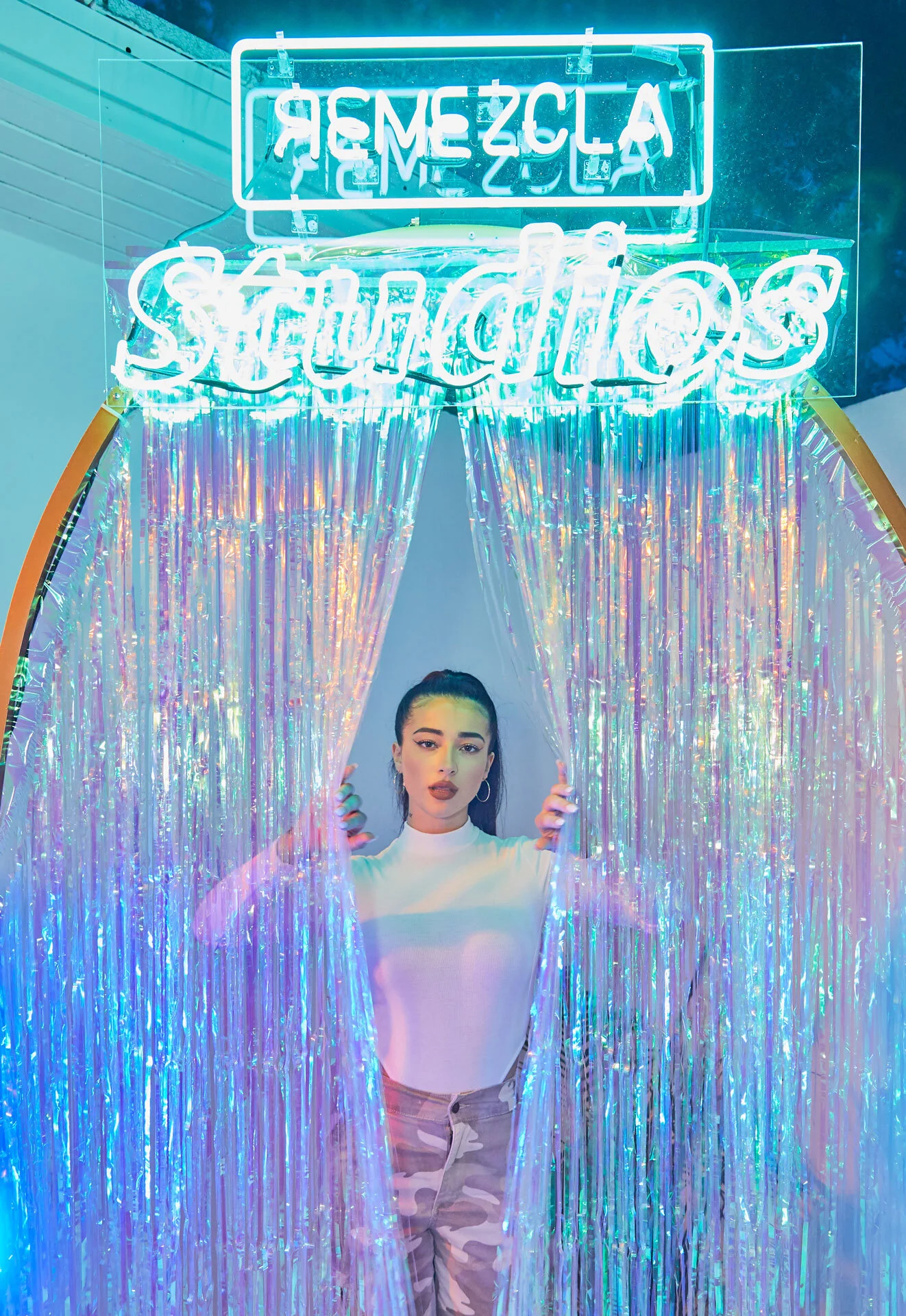 Joven mujer posando frente a cortina de hilos iridiscentes con cartel de neón que dice 'Reemzca' y 'Stand Up' en un fondo oscuro.