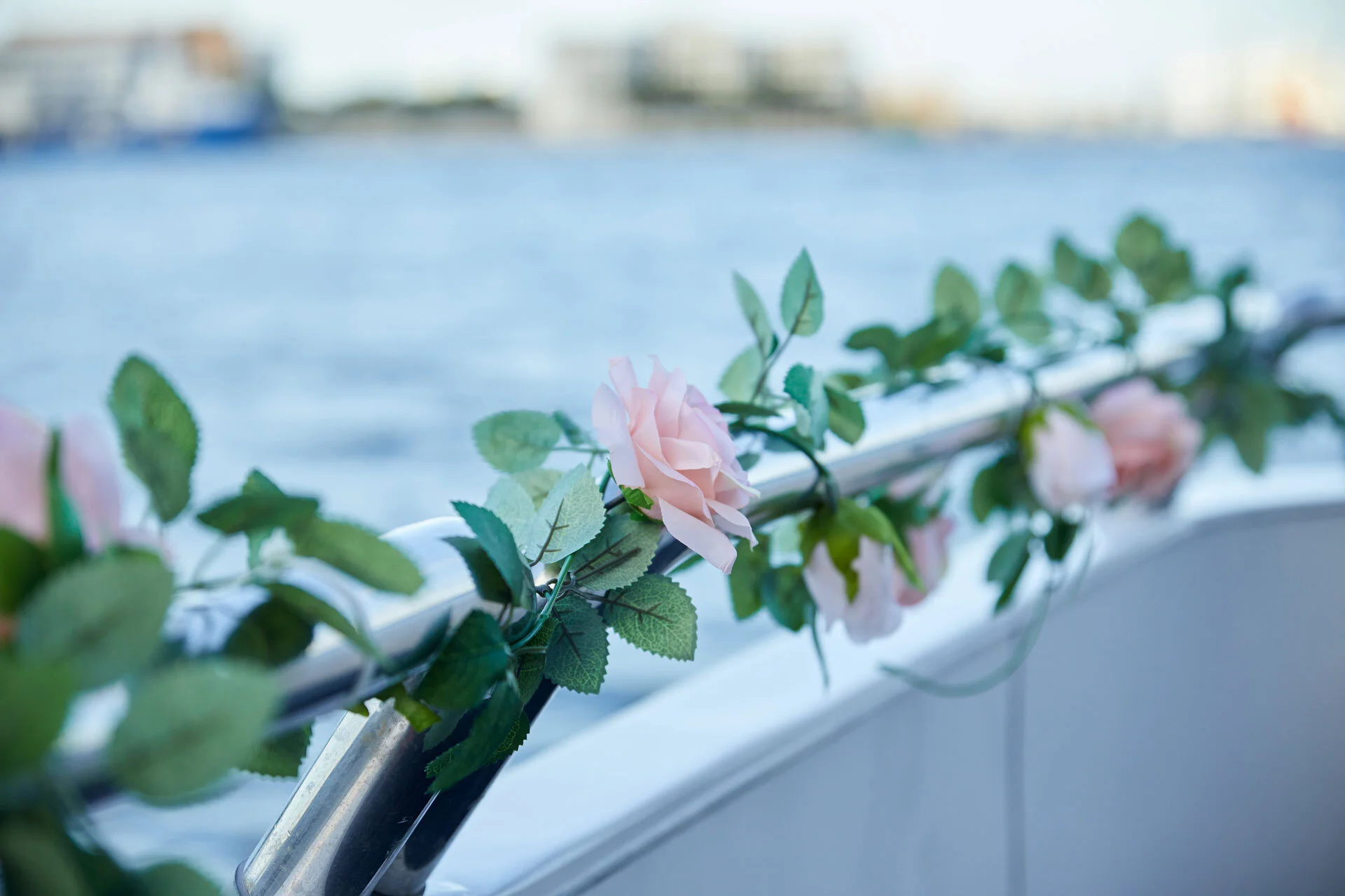 Decoración con rosas rosadas y hojas verdes en la barandilla de un barco, con fondo de agua y una ciudad borrosa.