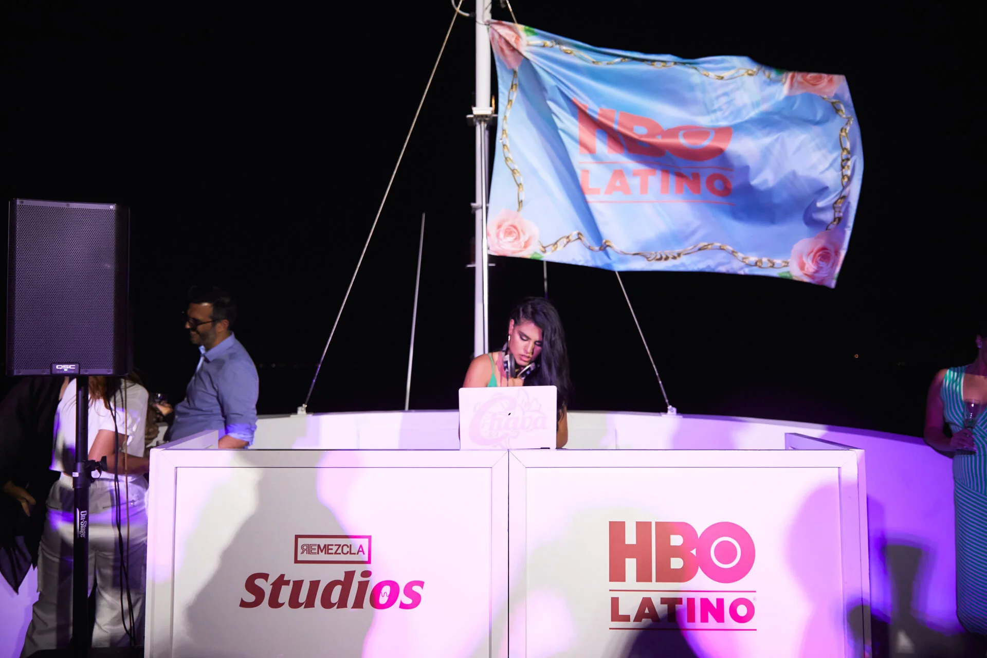 Una mujer en una cabina de DJ en un evento nocturno, con un cartel de HBO Latino y una bandera con el logo de HBO Latino en el fondo.