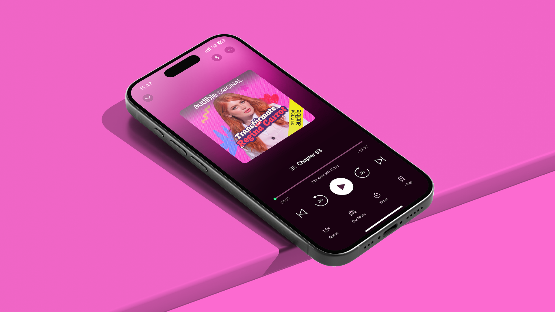 Un teléfono inteligente con pantalla que reproduce un audiolibro llamado 'Transfórmate' de Regina Catrone en la aplicación Audible, sobre un fondo rosa