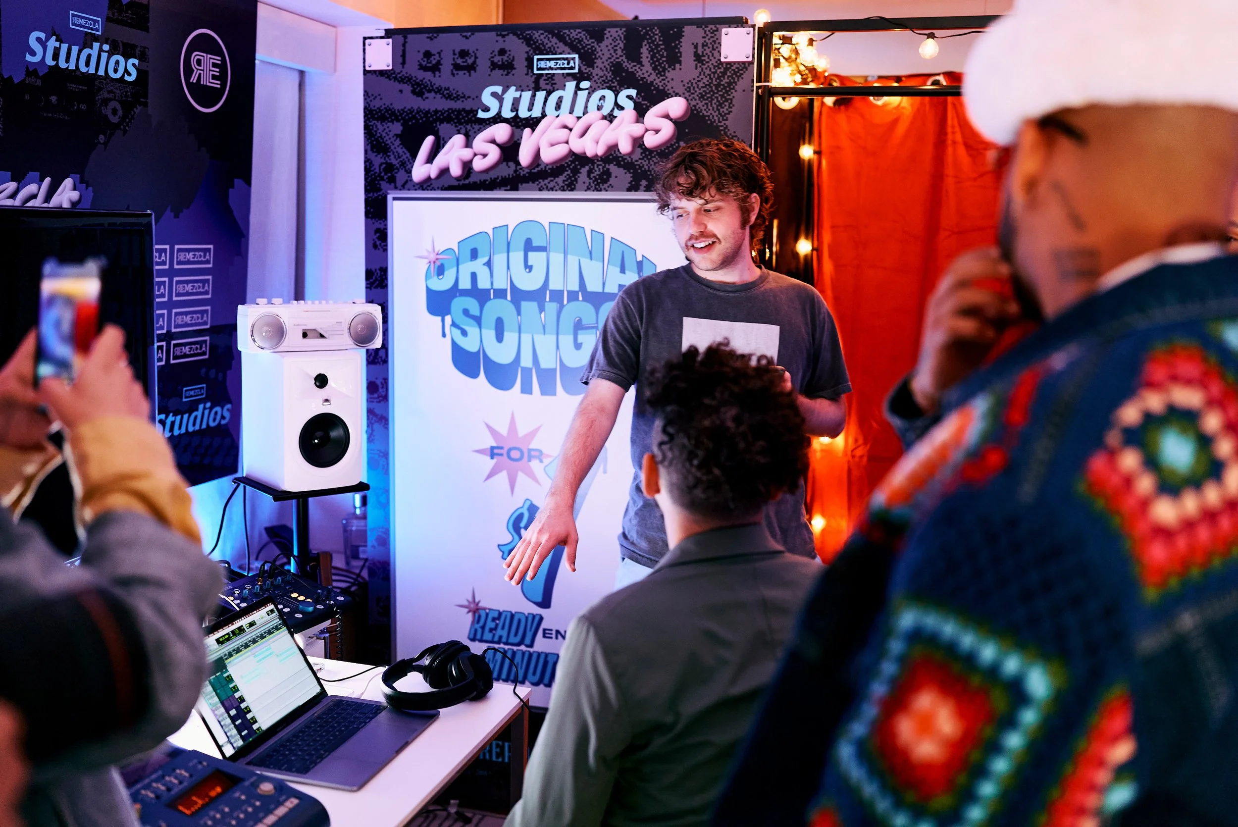 Un grupo de personas en un estudio de grabación con un presentador en el centro, mientras una persona toma una foto con su teléfono, en un ambiente decorado con luces y fondos con letras en inglés que dicen 'Estudios Las Vegas' y 'Original Song'.