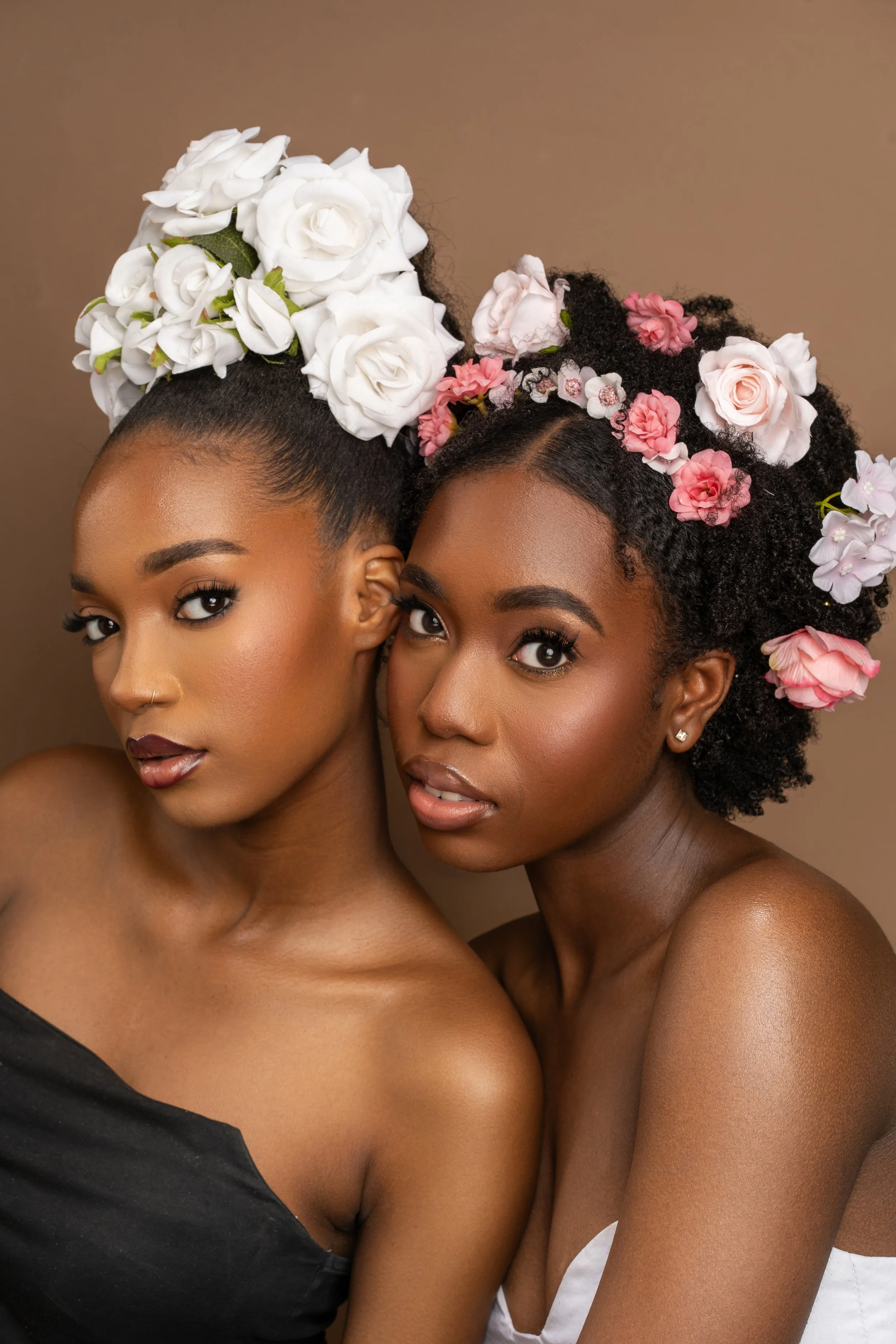 Two women with floral crowns posing closely for a portrait against a neutral background.