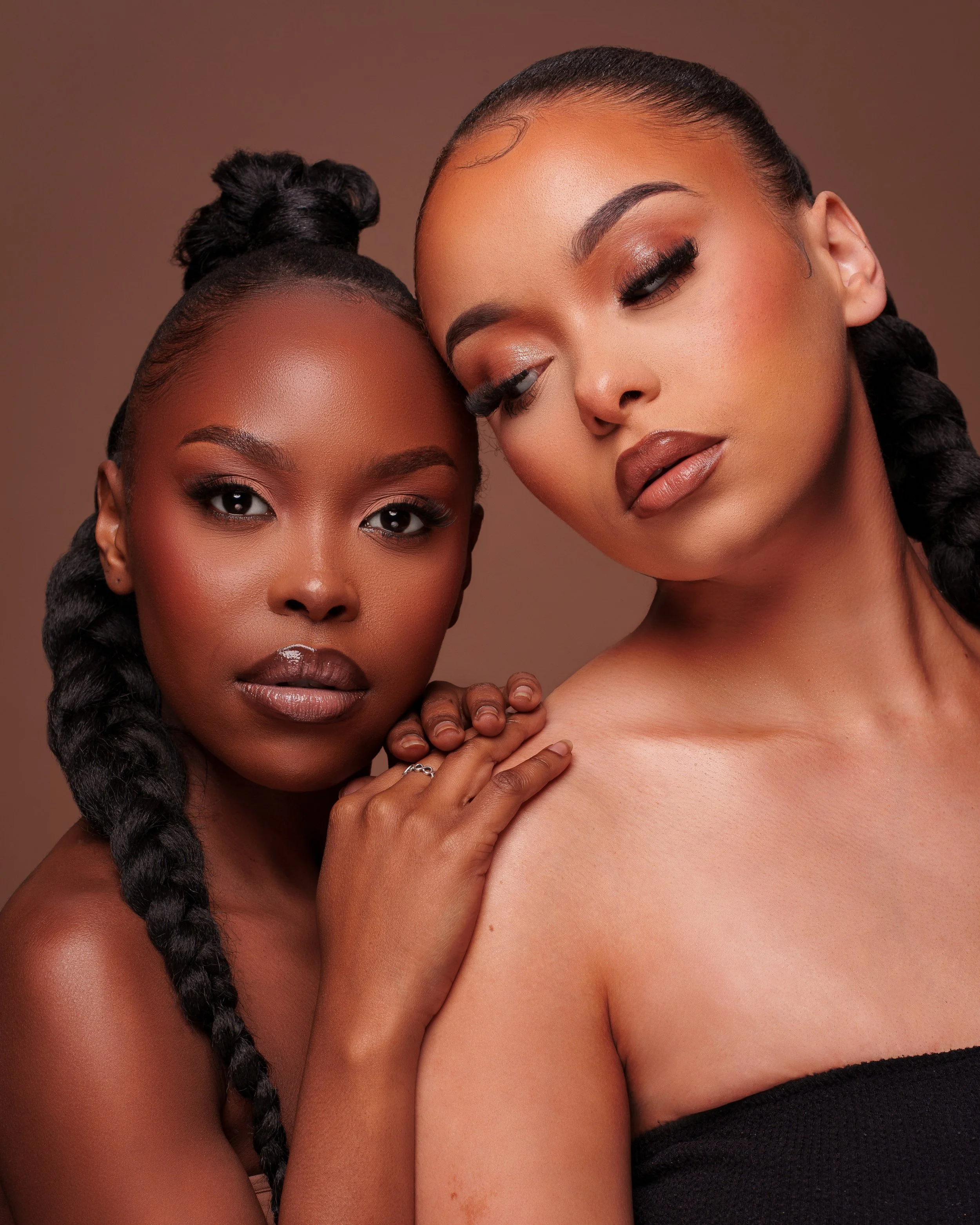 Two women with braided hair and makeup, one with eyes closed gently touching the other's shoulder, both looking at the camera, against a neutral background.