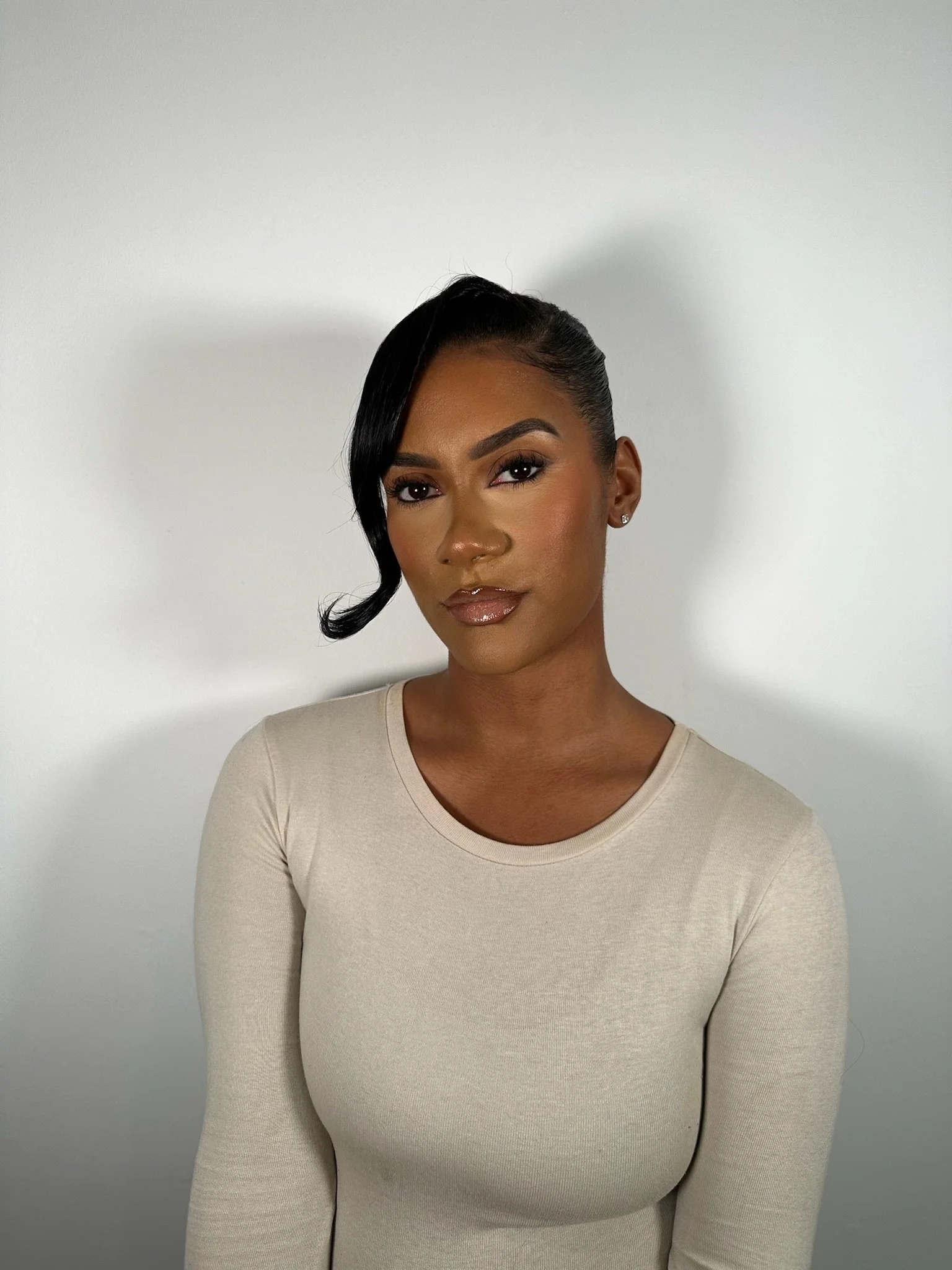 A woman with dark hair styled in an asymmetrical bob, wearing a beige long-sleeve top, standing against a plain white wall.