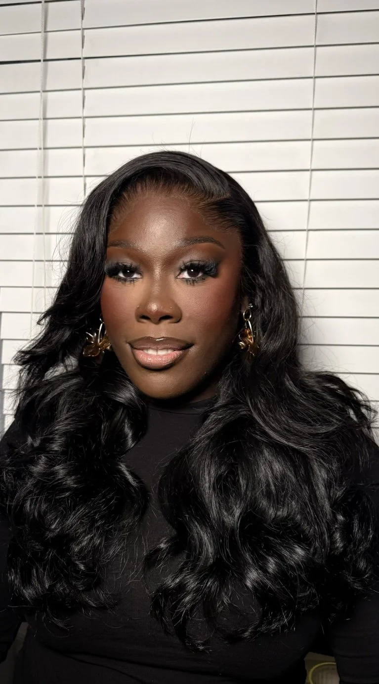 A woman with long, wavy black hair, wearing makeup and black clothing, standing in front of a white wall with horizontal blinds.