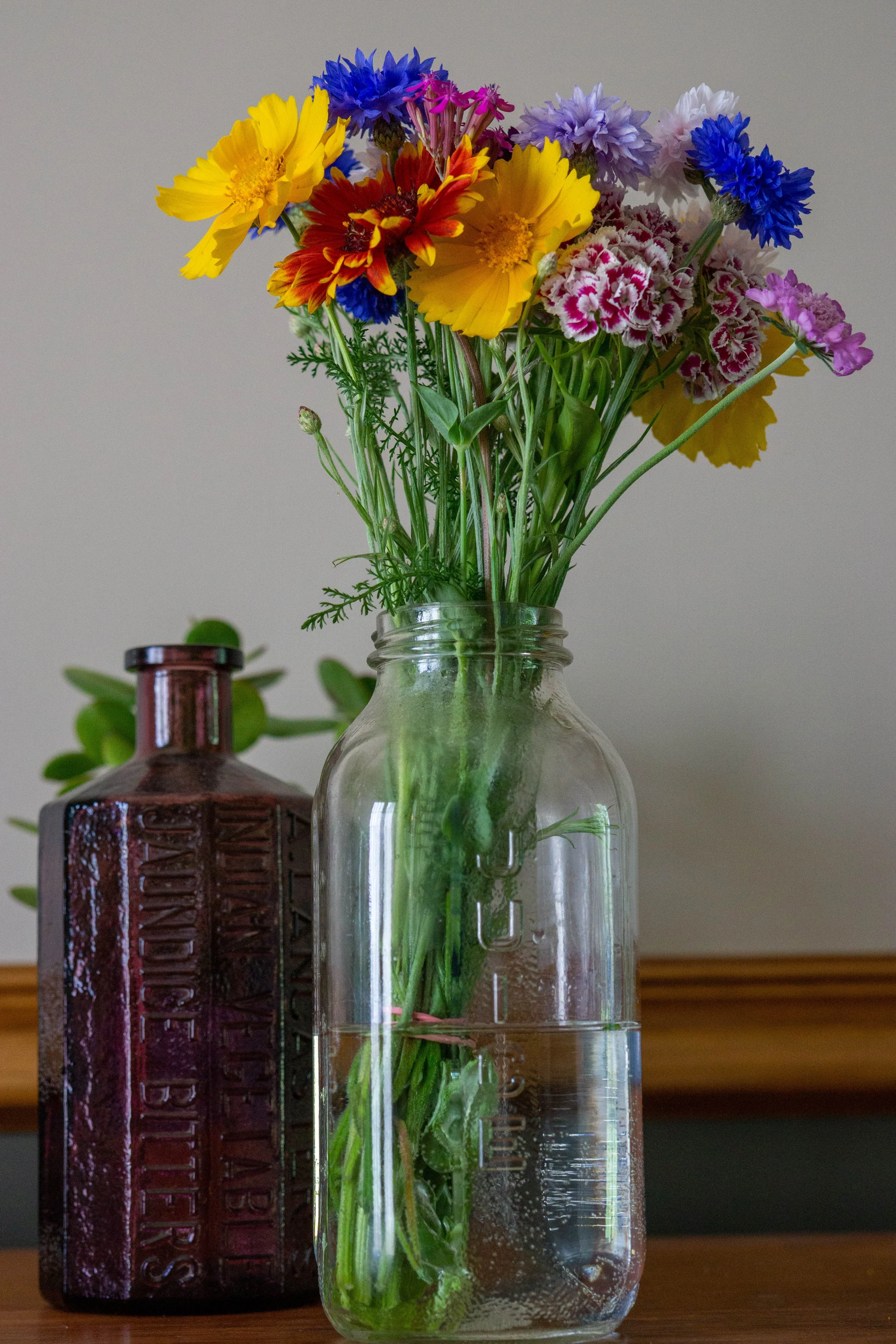 A clear glass jar filled with water and colorful flowers, including yellow, red, purple, and blue blooms, sits on a wooden surface next to a dark red bottle with embossed text.