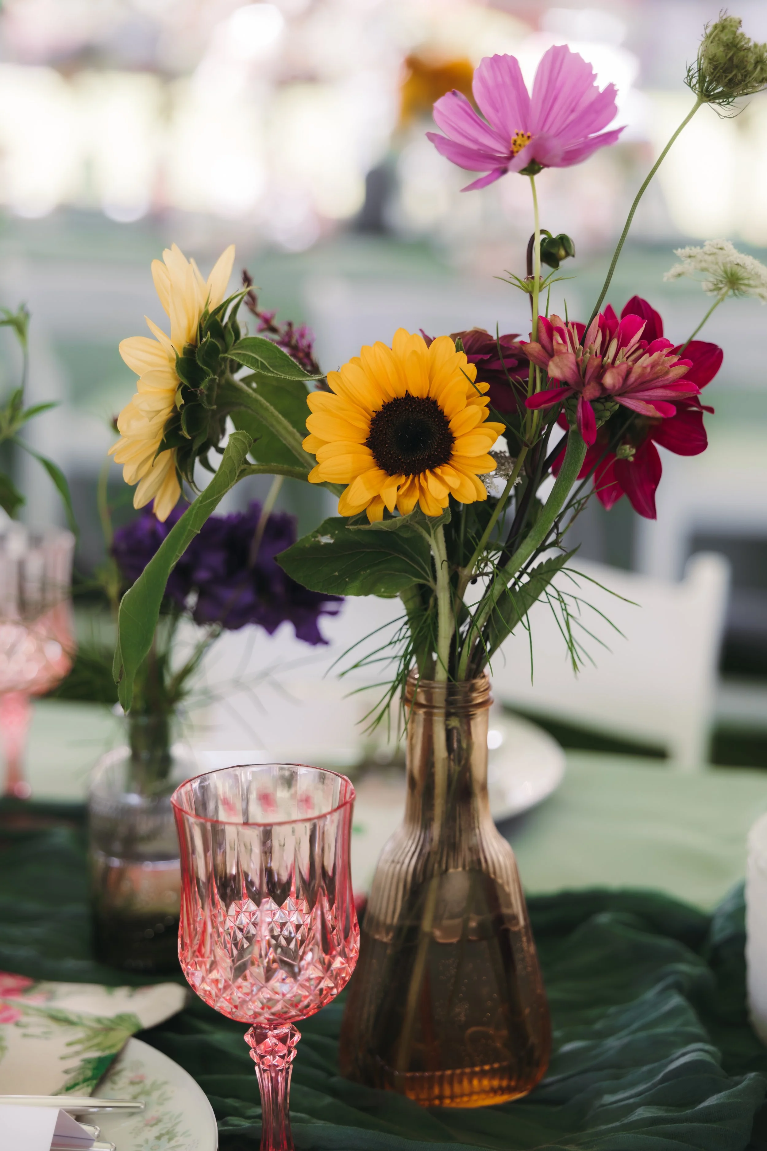 A bouquet of colorful flowers in a amber vase on a table with green tablecloth, with pink crystal glass and dinnerware, setting a festive or decorative scene.