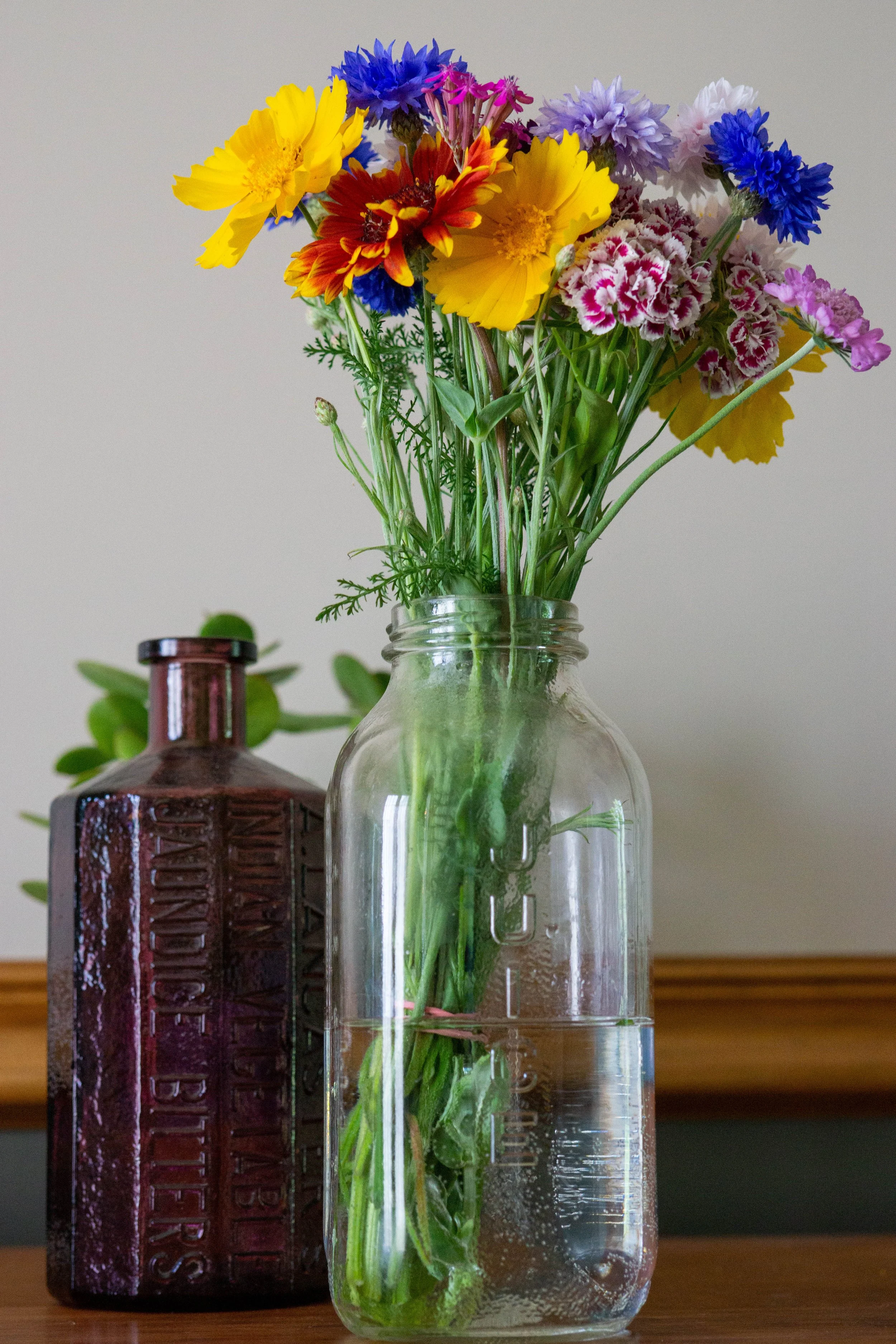A clear glass jar with water and green leafy stems holds a colorful bouquet of flowers including yellow, red, purple, pink, and blue blooms. A dark brown decorative bottle with embossed lettering stands behind the jar on a wooden surface against a pl