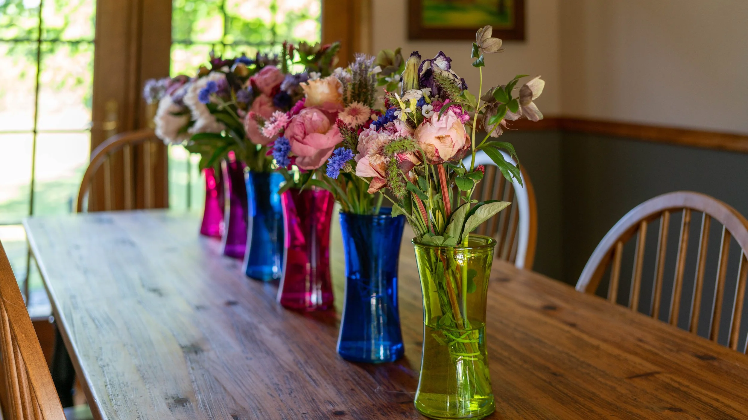 A wooden dining table with five colorful glass vases holding various pink, purple, and blue flowers, positioned near a window with natural light.
