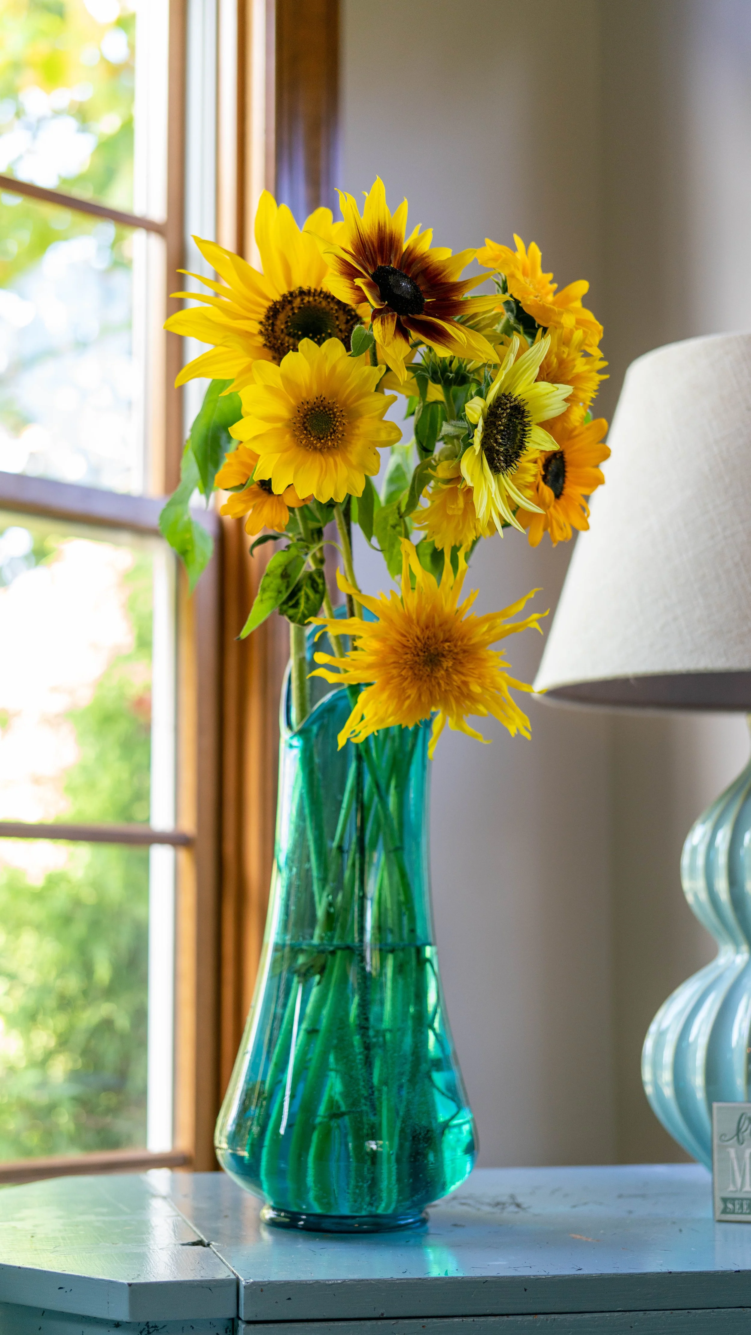 A bouquet of sunflowers in a green glass vase on a white wooden table near a window with sunlight.
