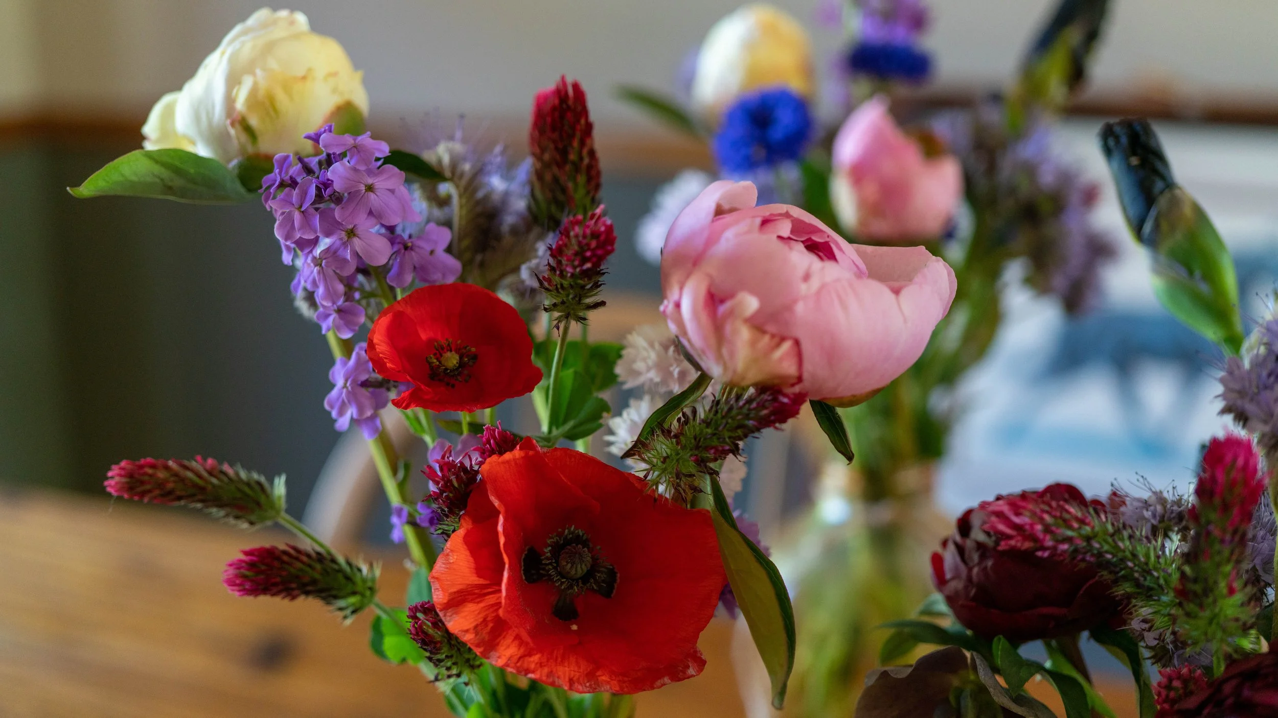 A colorful bouquet of various flowers including pink roses, red poppies, purple phlox, and other greenery in a vase on a wooden surface.