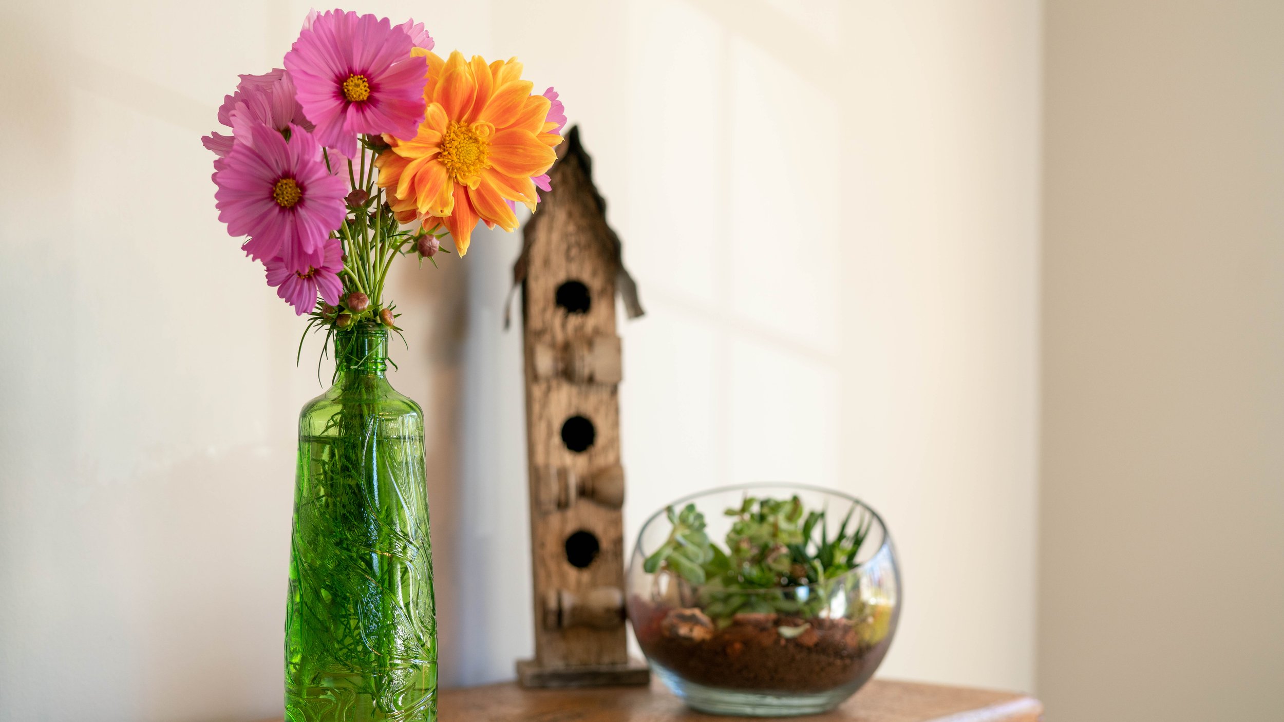A bouquet of pink and orange flowers in a green glass vase sitting on a wooden surface, with a wooden birdhouse and a glass bowl of succulent plants in the background.