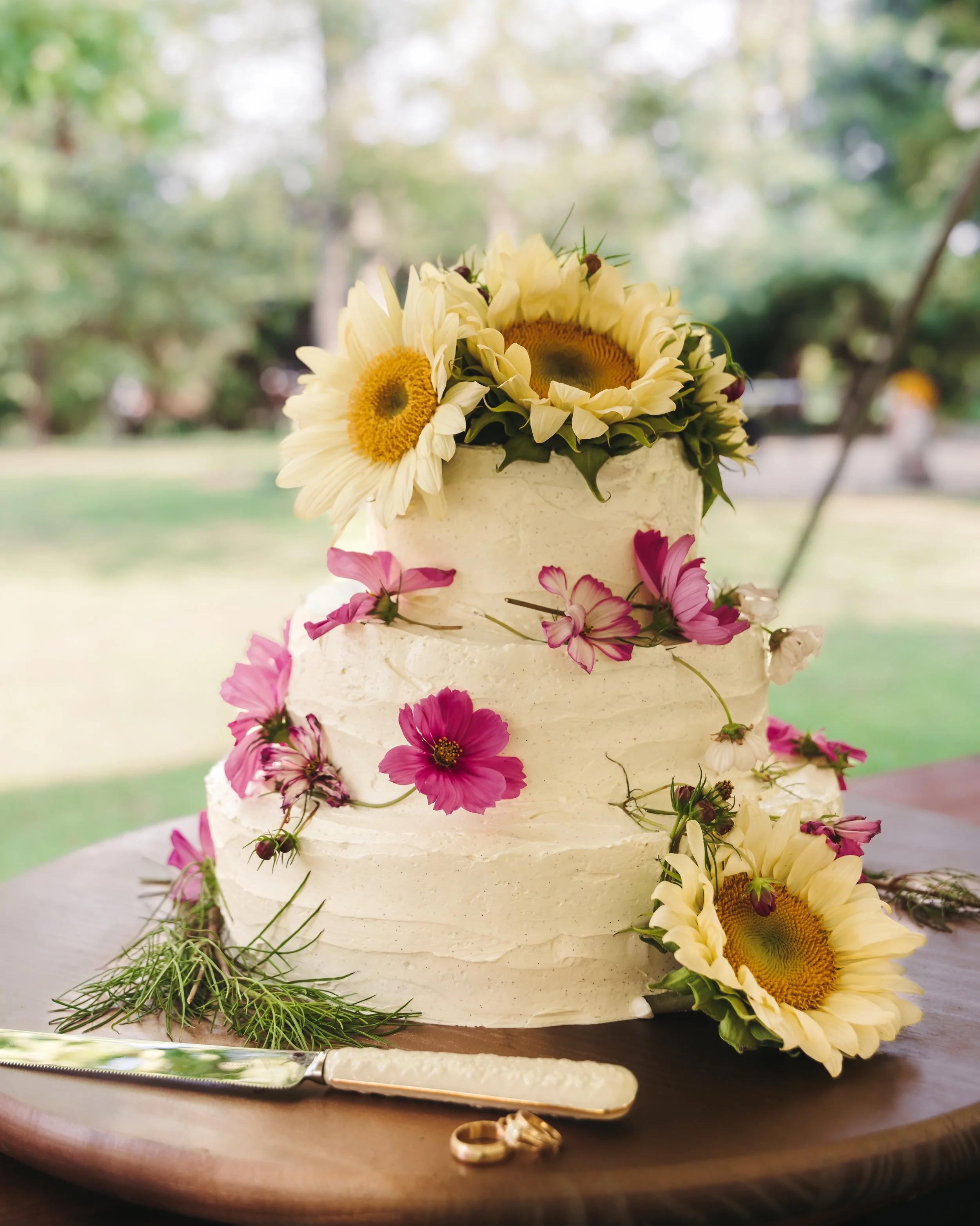A wedding cake decorated with sunflowers, pink and purple flowers, and green foliage, set on a wooden table outdoors.