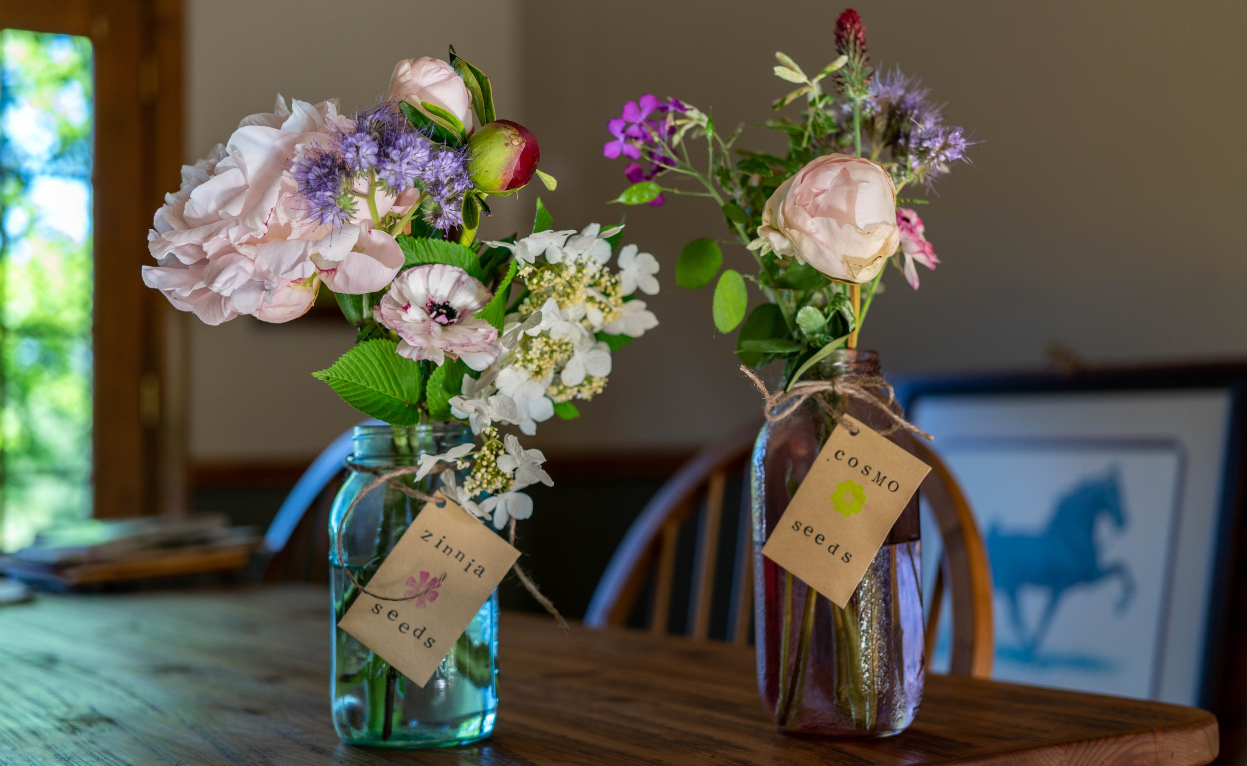 Two glass jars filled with various fresh flowers on a wooden table, with tags reading 'zinnia seeds' and 'cosmo seeds' attached to each jar, and a blurry background featuring a framed picture of a horse.