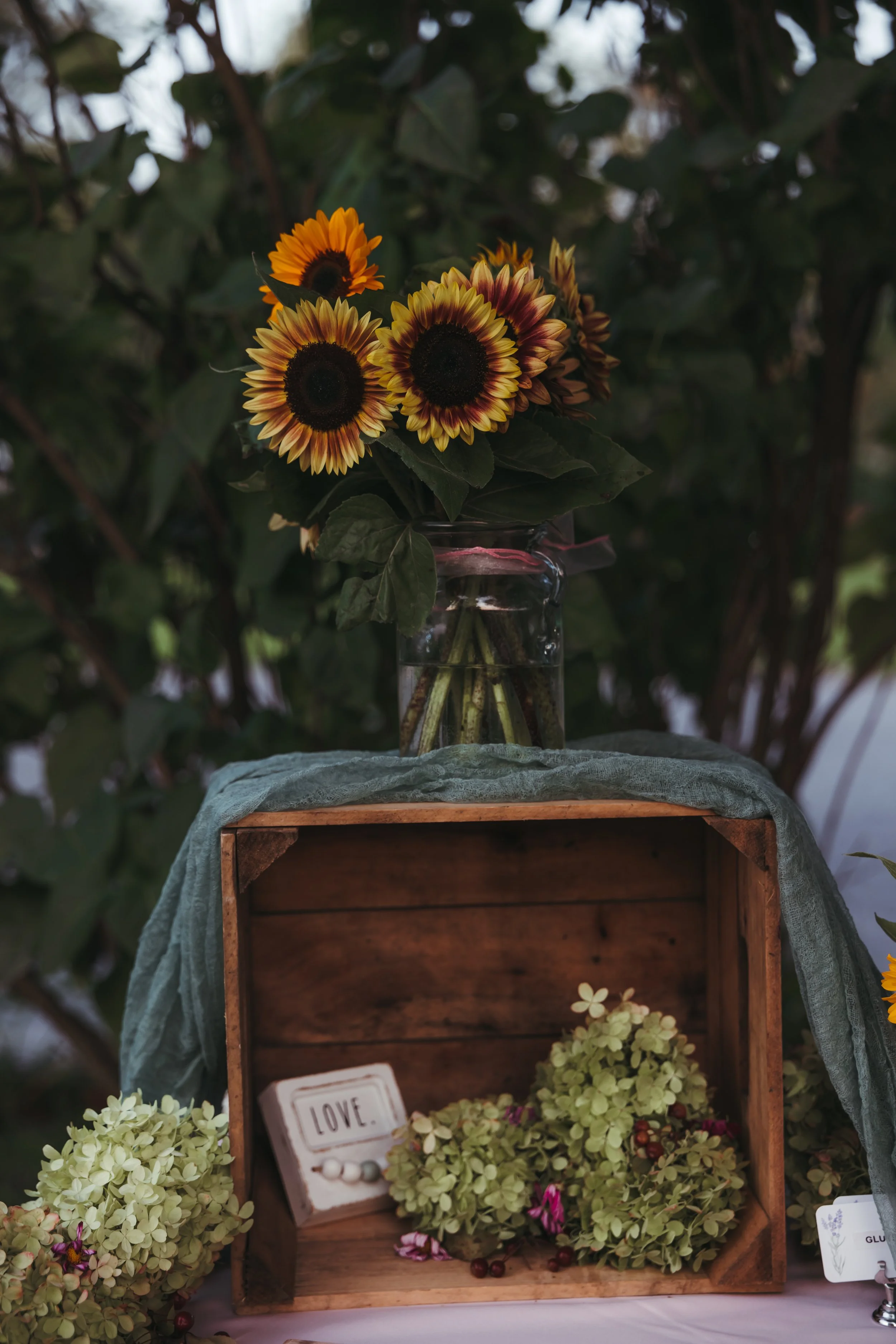 Vase of sunflowers on a table with a wooden crate filled with green hydrangeas and pink and purple flowers, with a small white sign that says 'LOVE' inside the crate.