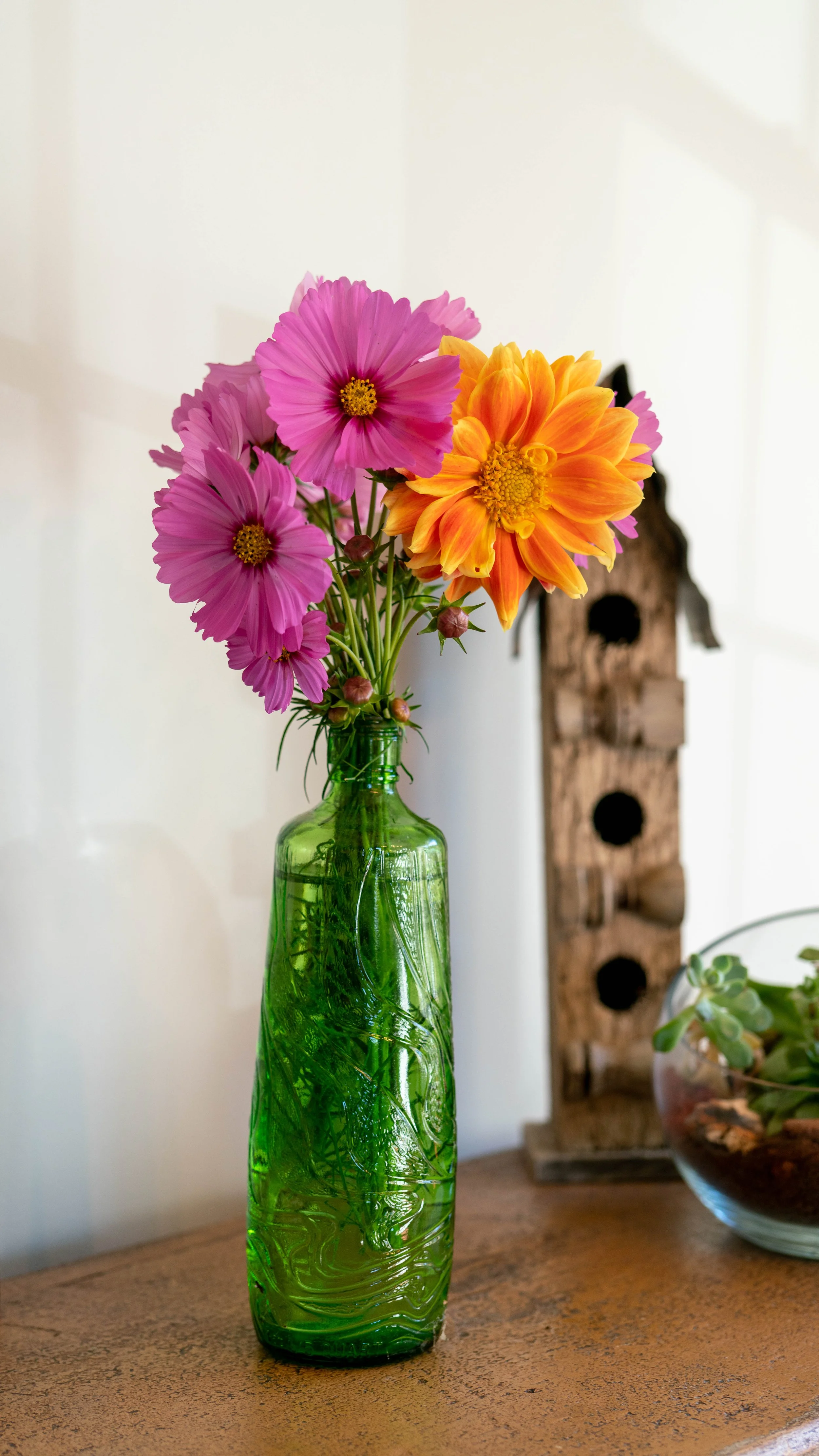 A green glass vase with pink and orange flowers on a wooden table, with a wooden birdhouse and a glass bowl with plants in the background.