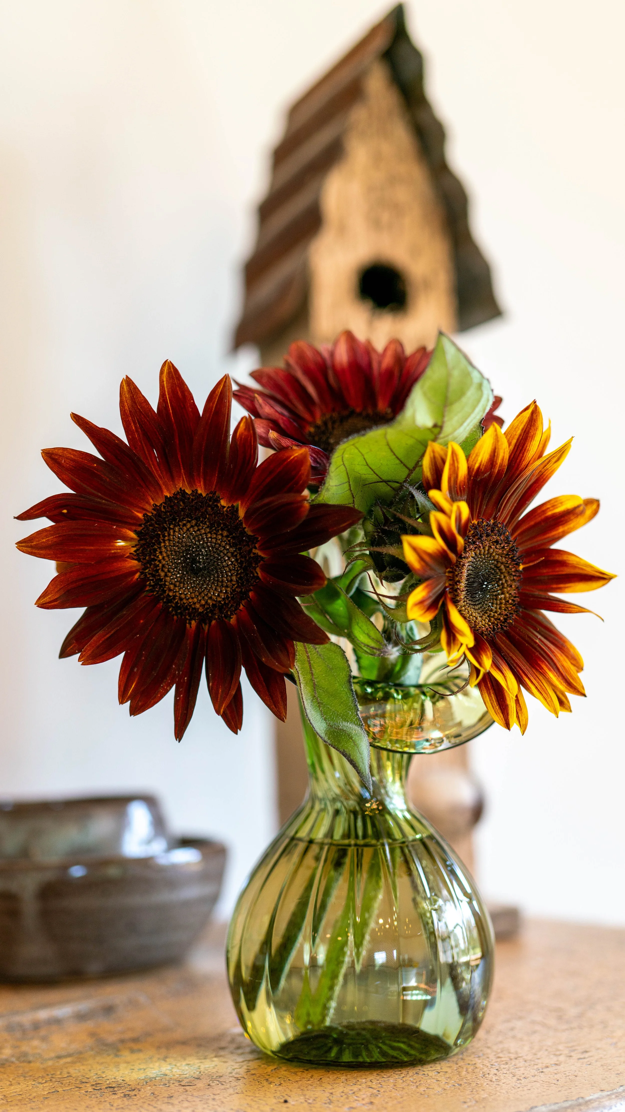 A bouquet of sunflowers with reddish-brown and orange petals in a clear glass vase on a wooden table, with a birdhouse in the background.