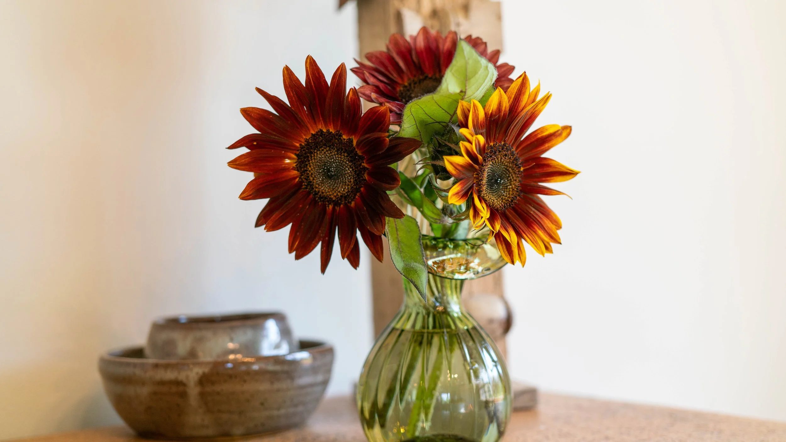 A glass vase containing sunflowers with reddish-brown and yellow petals, arranged on a wooden surface with a set of ceramic bowls in the background.