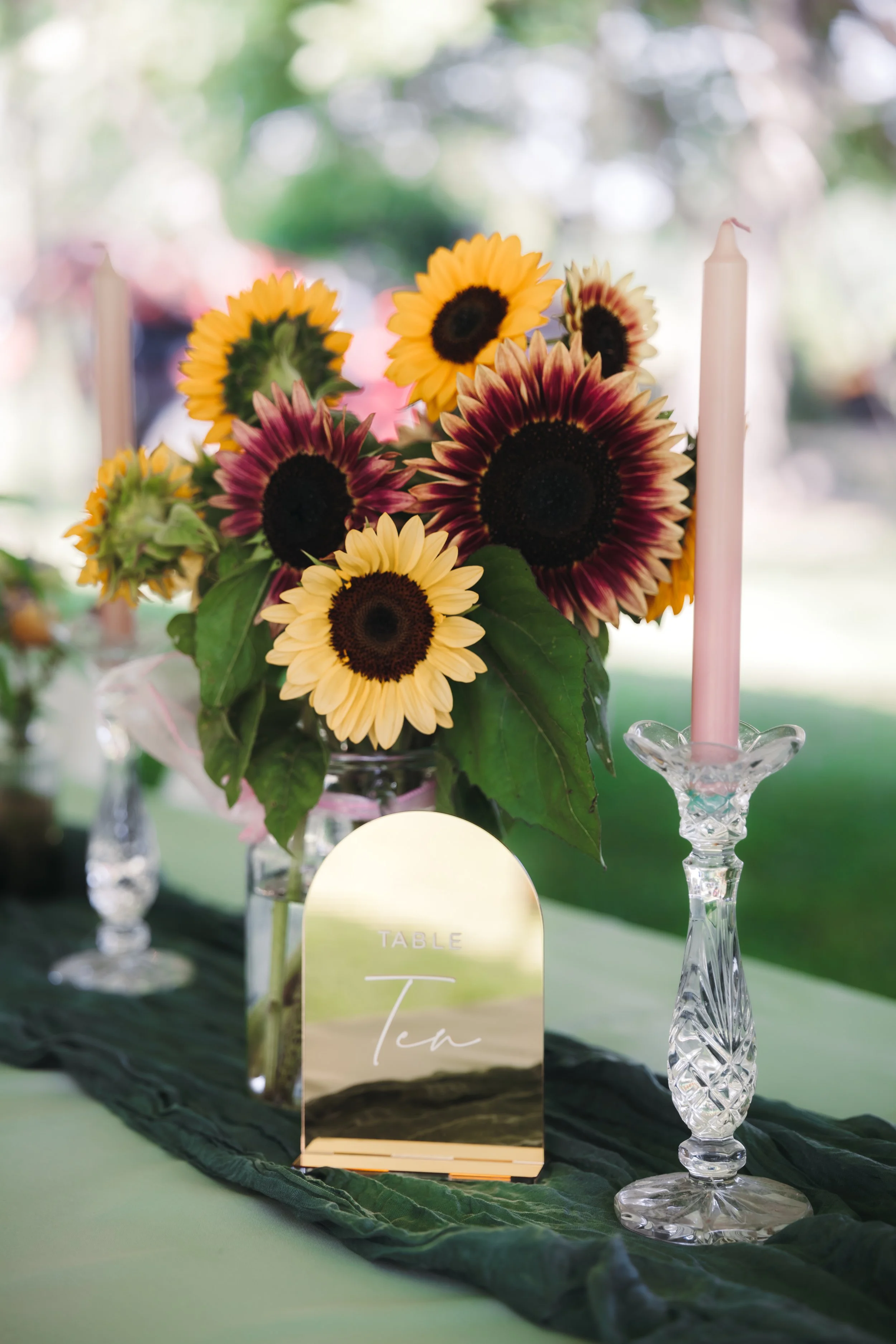 Arrangement of sunflowers and other flowers in a glass vase on a table with a black cloth runner, a pink candle in a crystal candlestick holder, and a gold-colored table sign that reads 'Table Tea'.