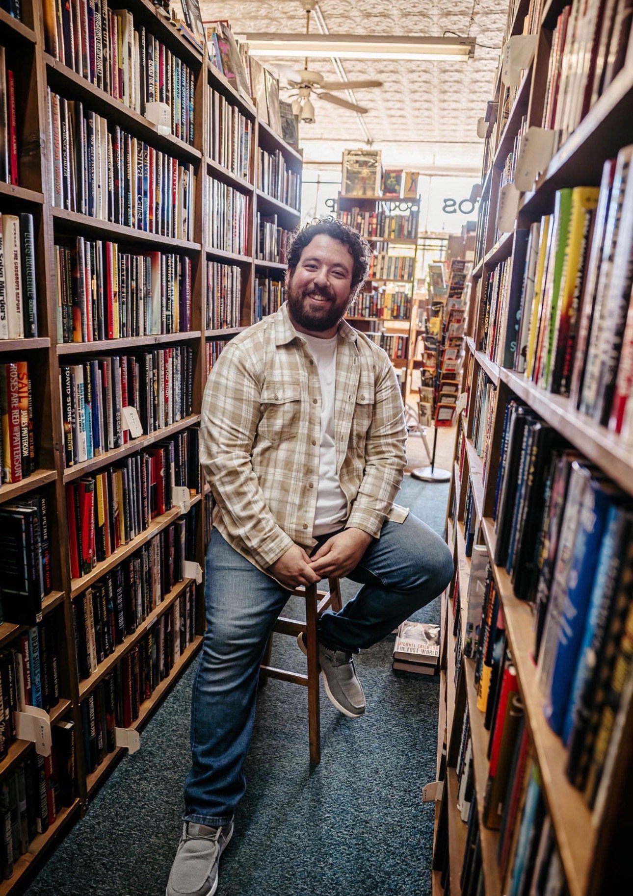 A man with a beard and curly hair smiling while sitting on a stool in a narrow aisle of a bookstore, surrounded by books on wooden shelves, with sunlight coming through windows in the background.