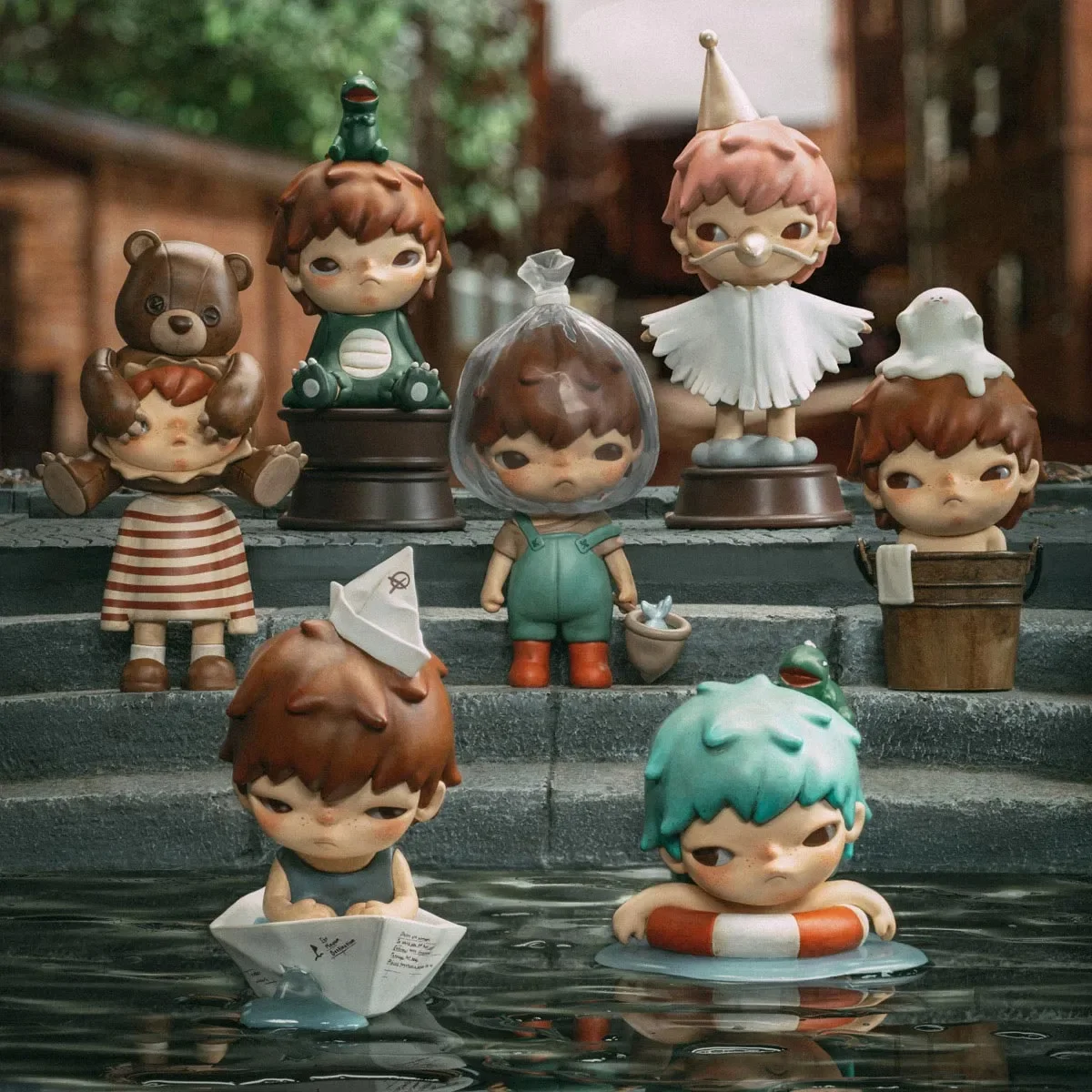 Collection of cute, cartoonish figurines with sad or neutral expressions, some with accessories like hats and a dog, placed on steps near water, with some in a small boat and others on pedestals.