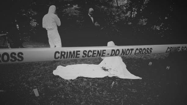 A crime scene with police tape reading 'CRIME SCENE - DO NOT CROSS' and a body covered by a white sheet, with investigators nearby in a wooded outdoor area.