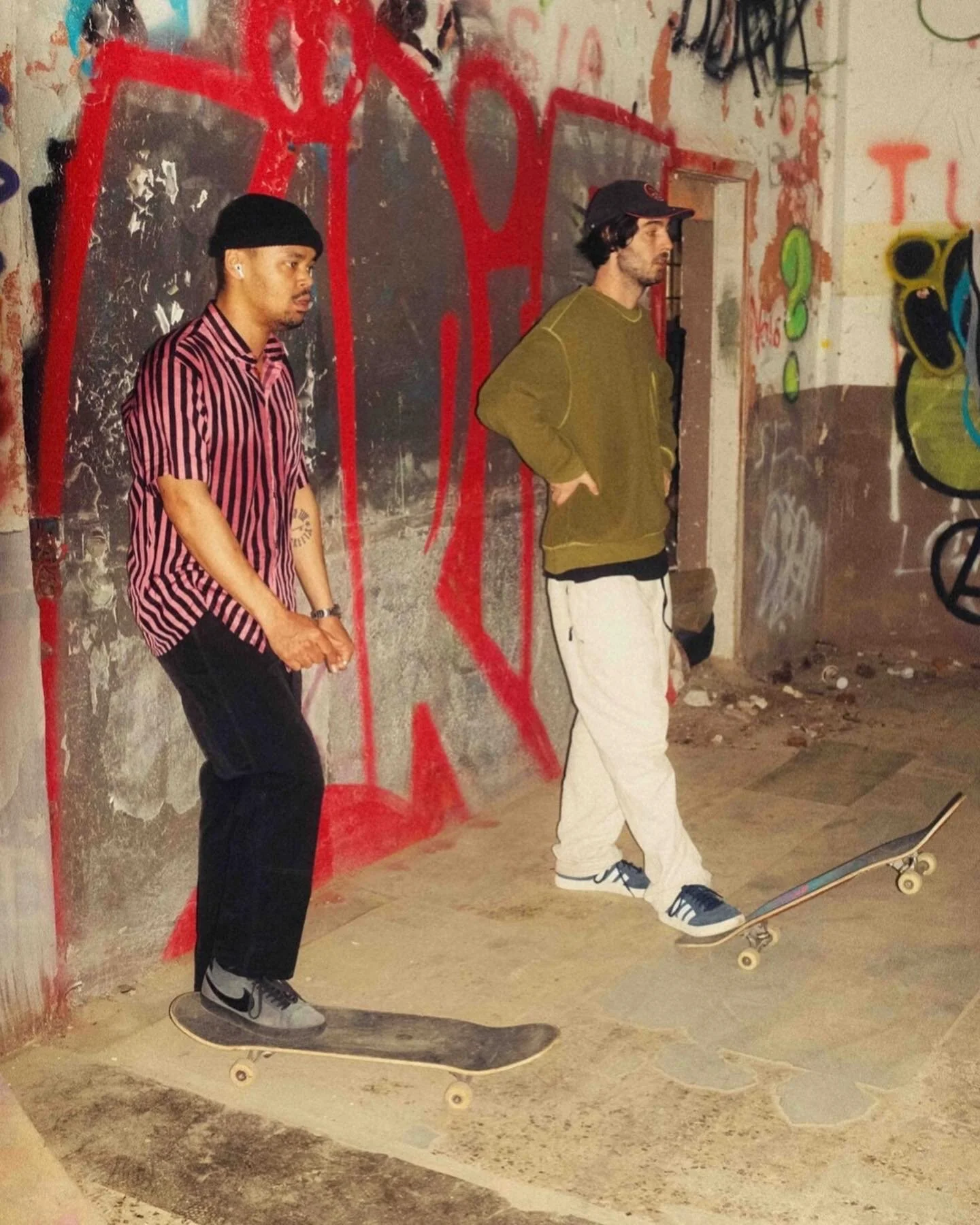 Zwei junge Männer springen auf Skateboards in einer graffiti-verzierten, dunklen und rauen Umgebung.