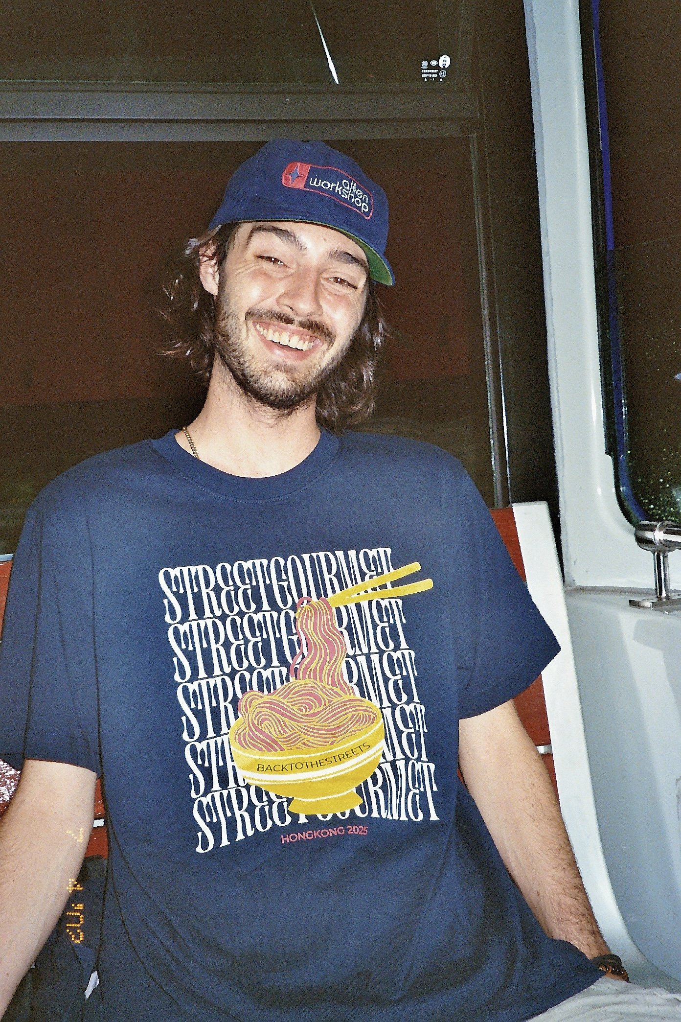 Ein lachender junger Mann trägt eine blaue Kappe und ein T-Shirt mit einem Nudel-Design. Er sitzt in einem Fahrzeug, wahrscheinlich einem Bus, während es regnet.