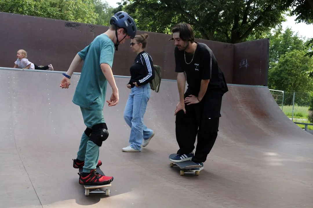 Zwei Jugendliche üben Skateboardfahren auf einer Halfpipe, während ein dritter Teenager zuschaut. Im Hintergrund sitzt ein kleines Mädchen auf der Randkante der Halfpipe.