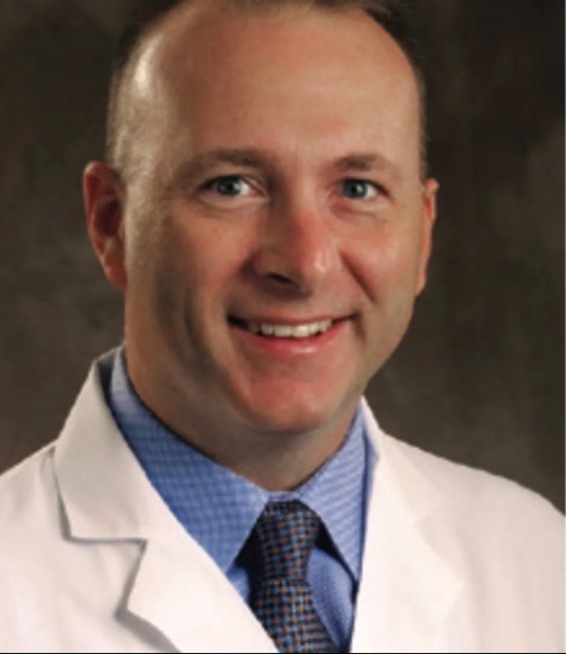 A smiling man in a white medical coat and a blue shirt with a patterned tie, posed in a professional portrait setting.