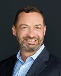 Professional headshot of a middle-aged man with short dark hair and a beard, smiling, wearing a dark blazer and light blue shirt against a dark background.