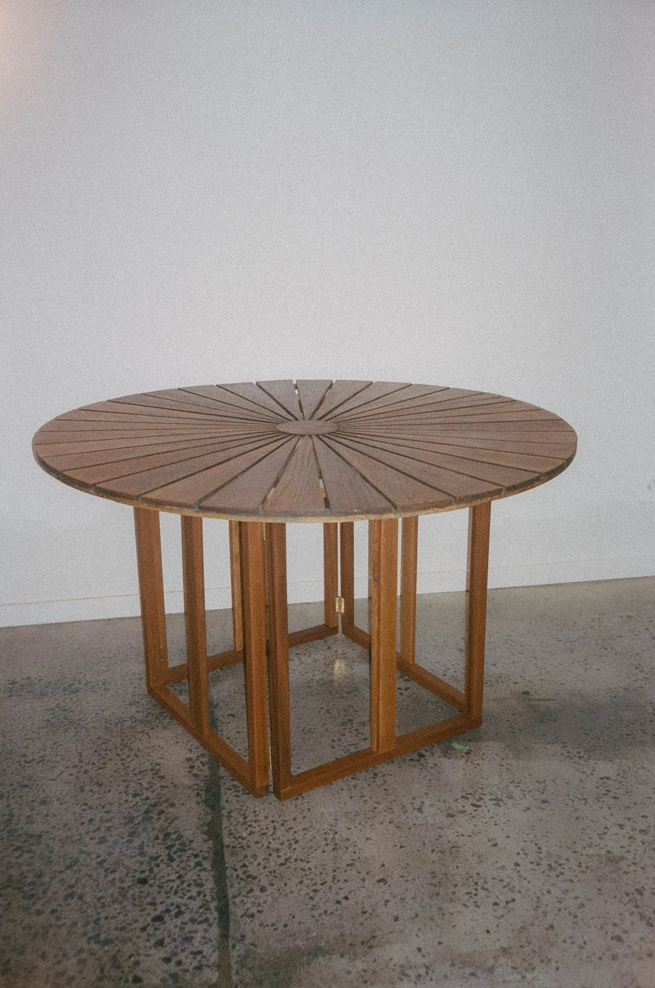 A round wooden table with radial slats on top, supported by a square wooden frame with open sides, placed on a concrete floor against a plain white wall.