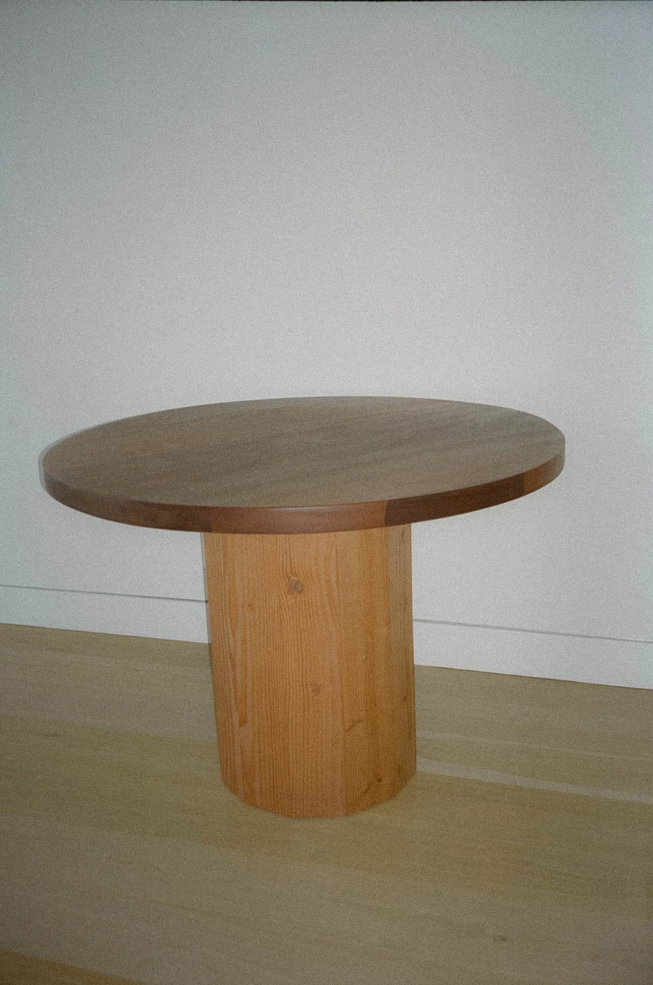 A wooden dining table with a round top and cylindrical base, placed against a white wall on a light wood floor.