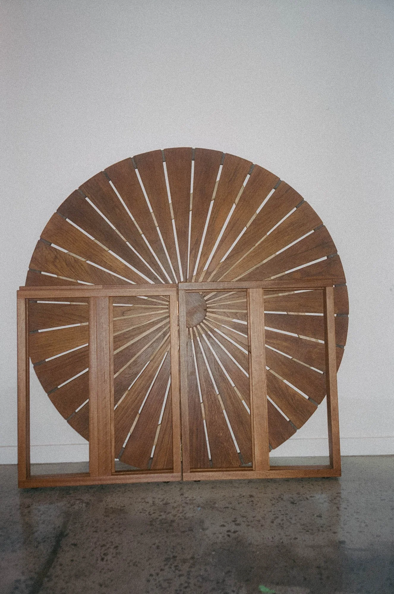 A large, curved wooden piece with radiating slats, leaning against a plain white wall and foldable a wooden framed base.