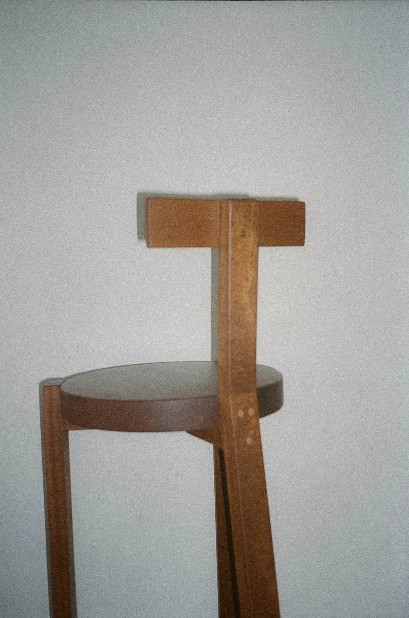 Close-up of a wooden stool with a rounded seat and a tall backrest against a plain wall.