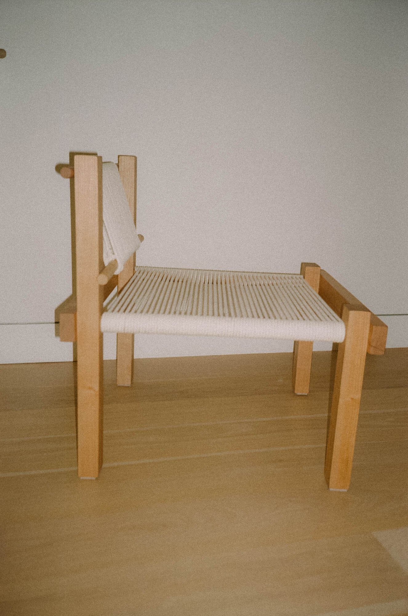 A wooden lounge chair with a woven seat and a slatted backrest, placed against a white wall on a wooden floor.