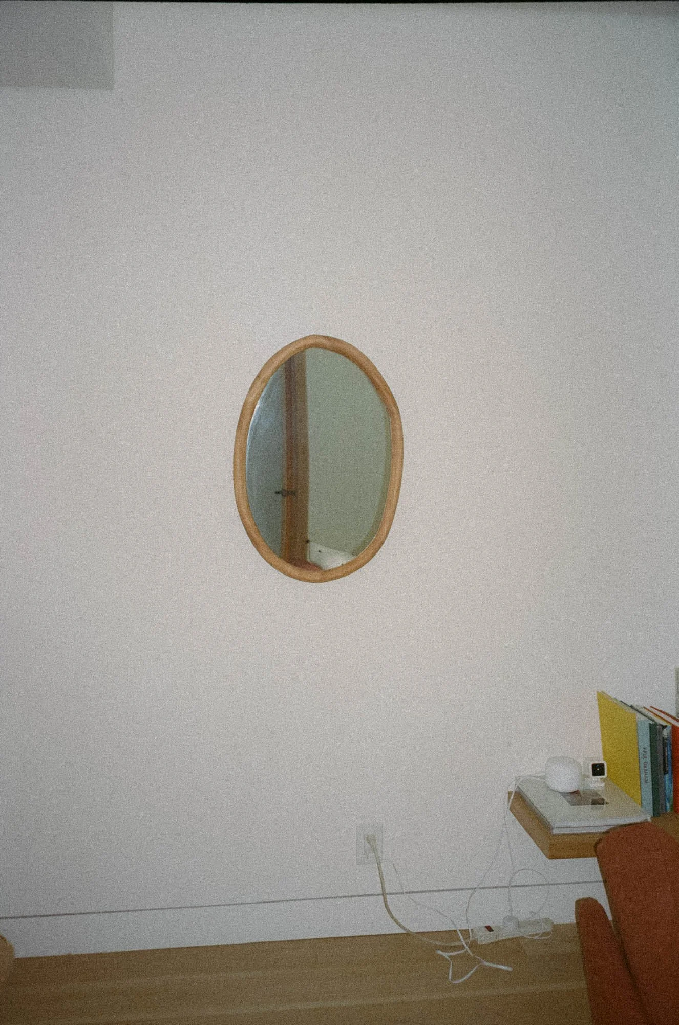 A white wall with an oval mirror and a small wooden shelf holding books, a white smart speaker, and a yellow file folder. An orange chair is partially visible in the lower right corner.