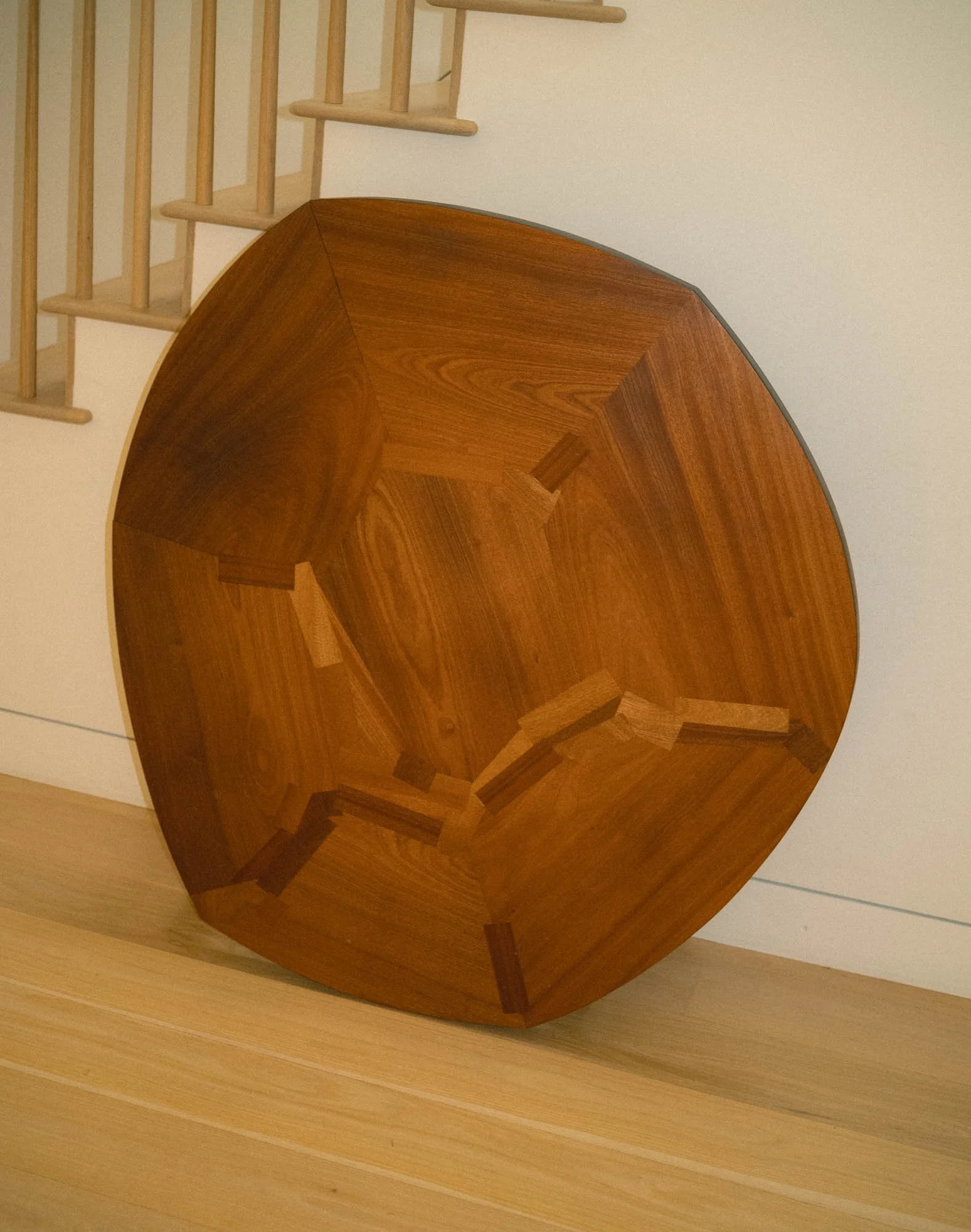 A organic shaped round wooden table with a geometric inlay pattern on the top, leaning against a white wall near a wooden staircase.