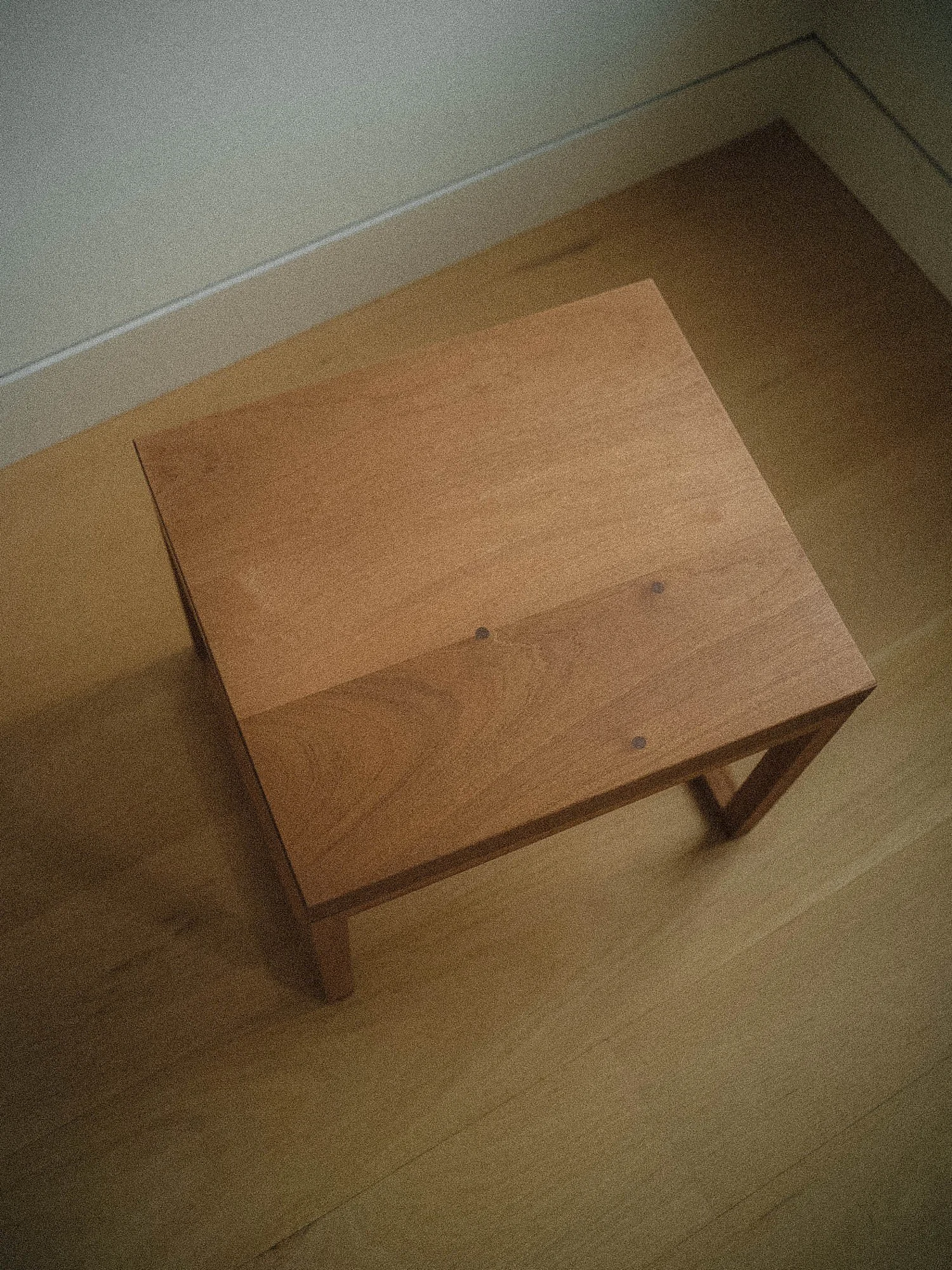 A wooden table with a square top, viewed from above, standing on a wooden floor next to a white wall.
