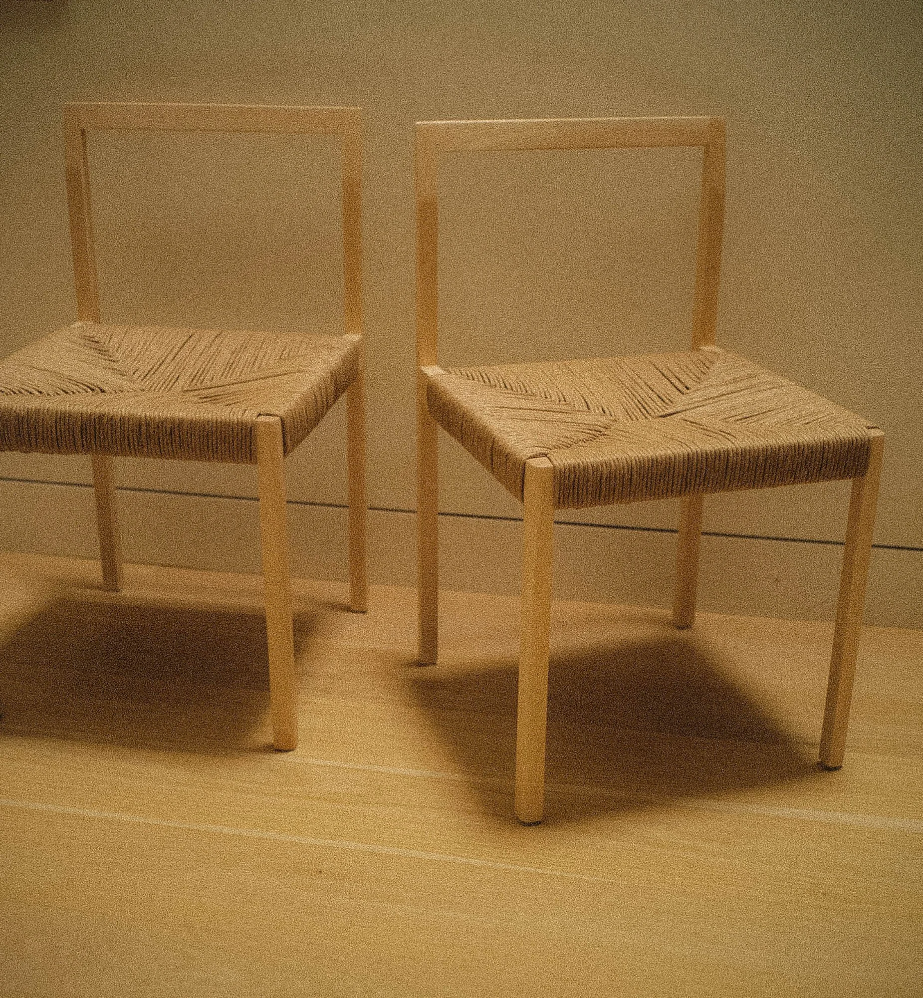 Two minimalist chairs with wooden frames and woven seats are positioned side by side in a plain room with beige walls and floor.