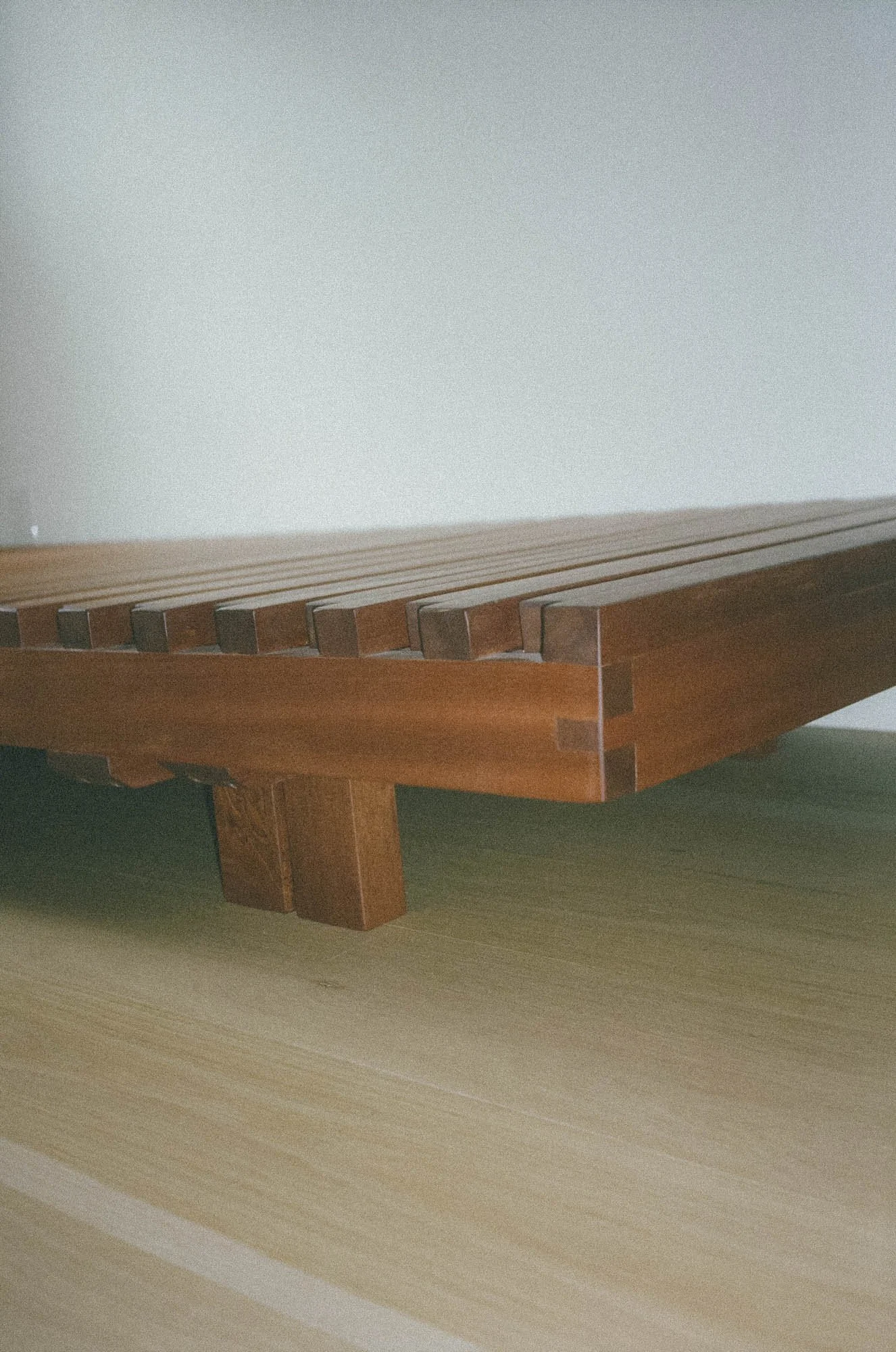 Low-angle view of a wooden platform or bed with side slats on a light-colored wooden floor against a plain wall.