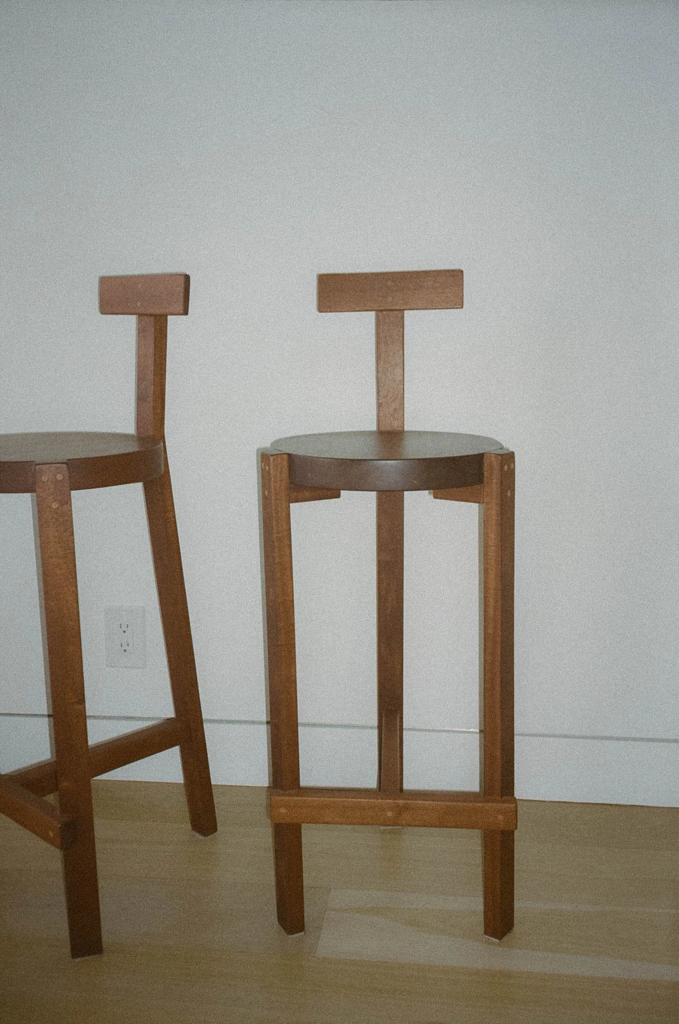 Two wooden chairs with simple design and no cushions, one partially visible on the left and one fully visible on the right, against a plain white wall with an electrical outlet near the bottom.
