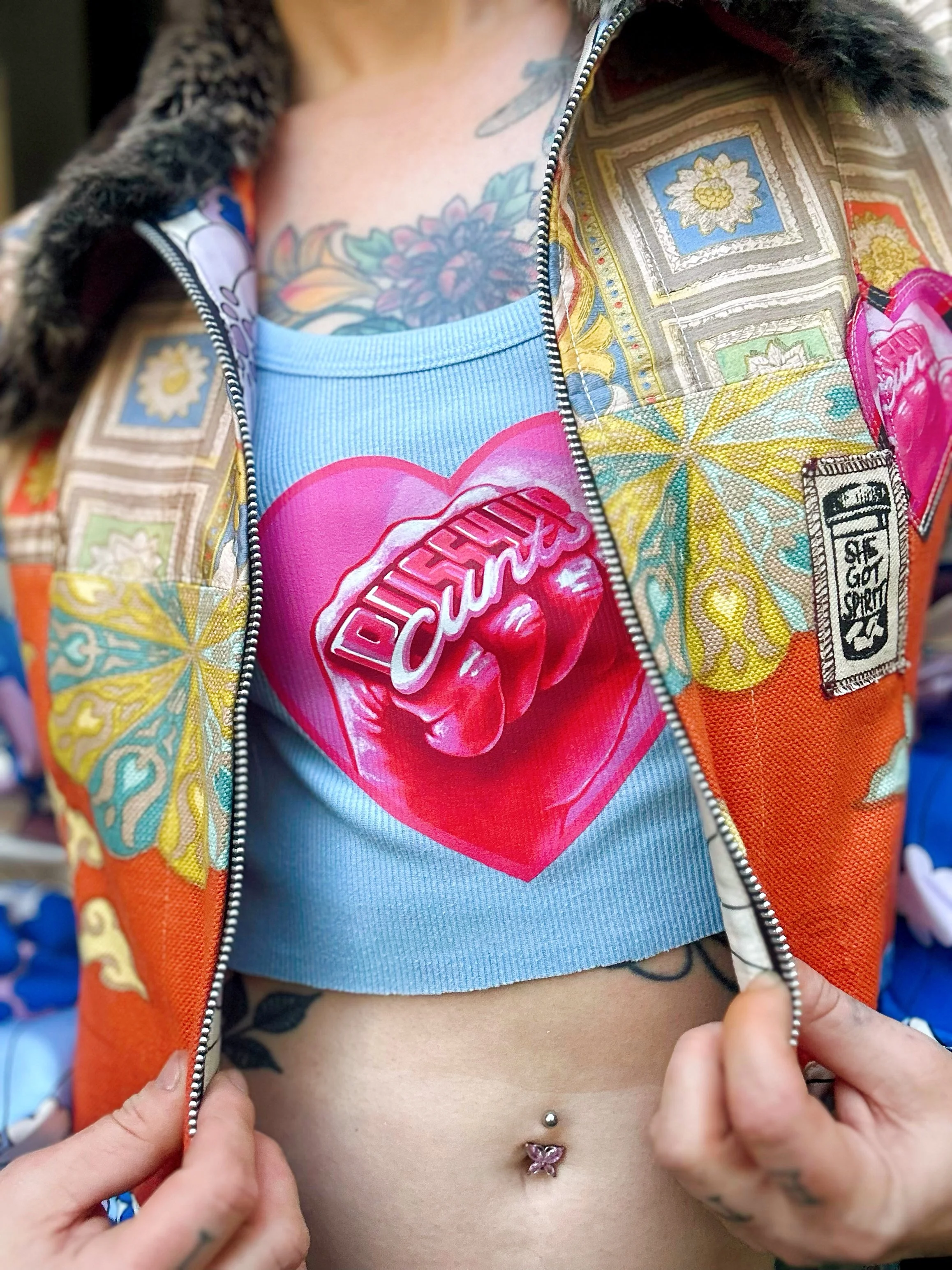 Close-up of a person revealing part of a colorful patterned jacket, a blue crop top with a pink heart and the words 'Dirty Souls,' a tattooed chest, and a navel piercing.