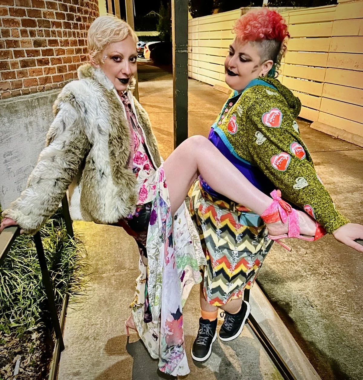 Two women with colorful hairstyles and bold makeup pose outdoors at night. One is holding the other up, revealing their legs. Both women wear vibrant, patterned clothing and high-heeled shoes. The woman on the left has platinum blonde hair and a faux fur coat, while the woman on the right has a rainbow-colored mohawk and wears a patchwork jacket. They appear playful and confident.