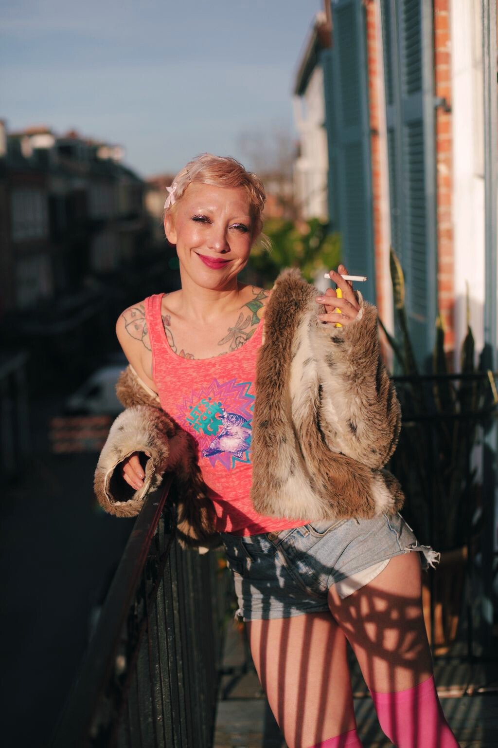 A woman with short blonde hair, tattoos on her shoulders, wearing a pink tank top and frayed denim shorts, stands on a balcony in sunlight, holding a cigarette and smiling.