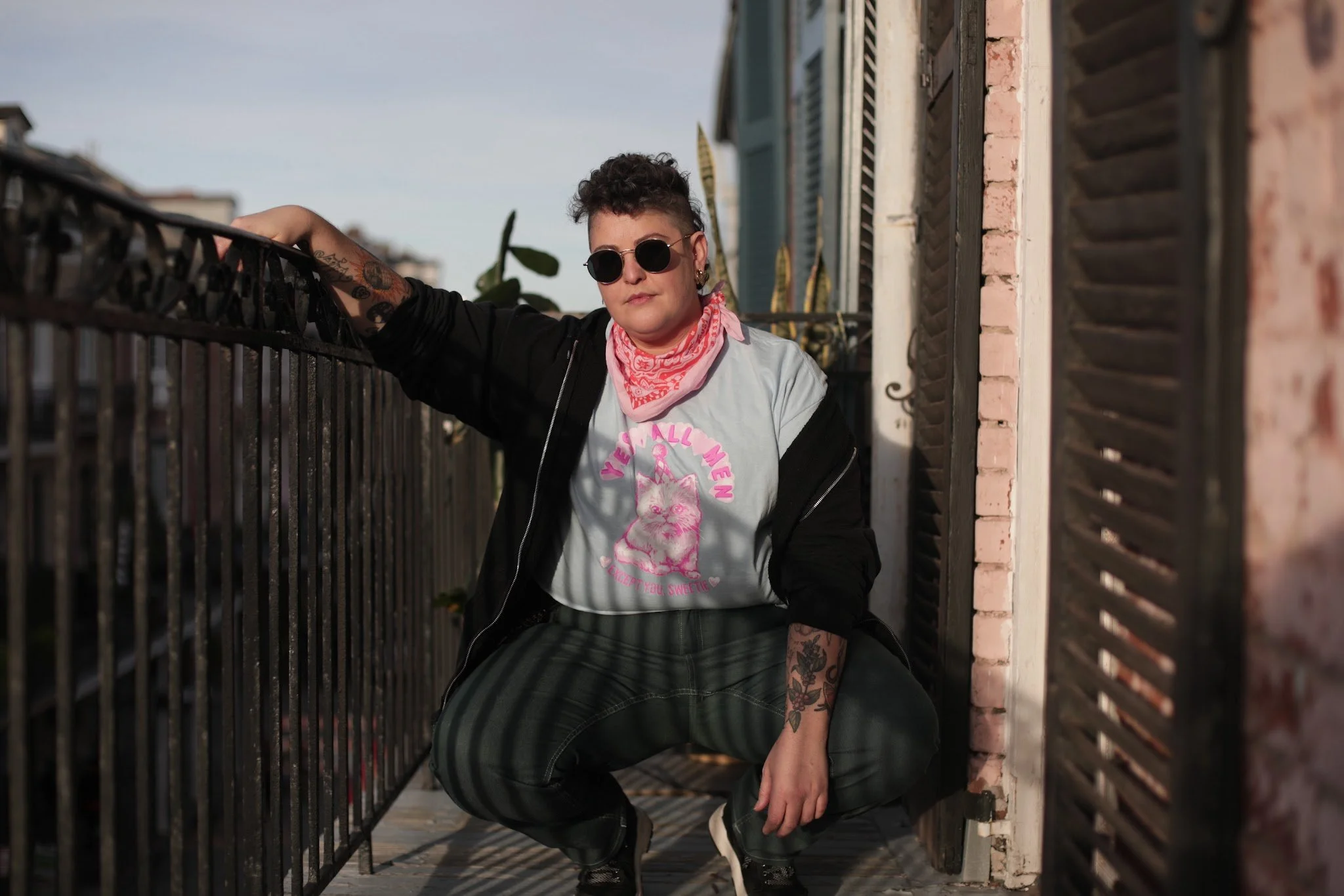 Person squatting on a balcony with a black metal railing, wearing sunglasses, a pink bandana, a graphic t-shirt with a cat and the words 'Very Luvly Wren,' a black jacket, striped pants, and sneakers, with tattoos visible on their arms, and buildings in the background.