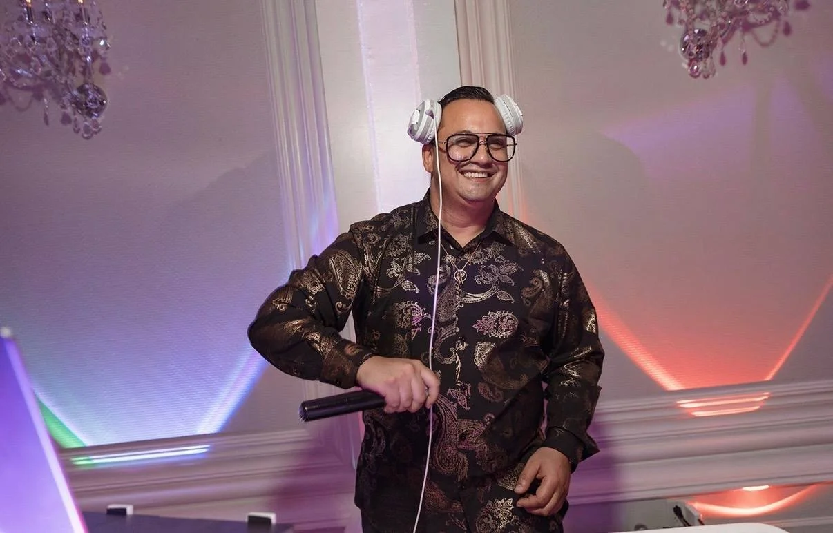 DJ Frankie G, smiling, wearing a decorative, dark patterned shirt, holding a microphone, and wearing headphones, at a party or event with colorful lighting.