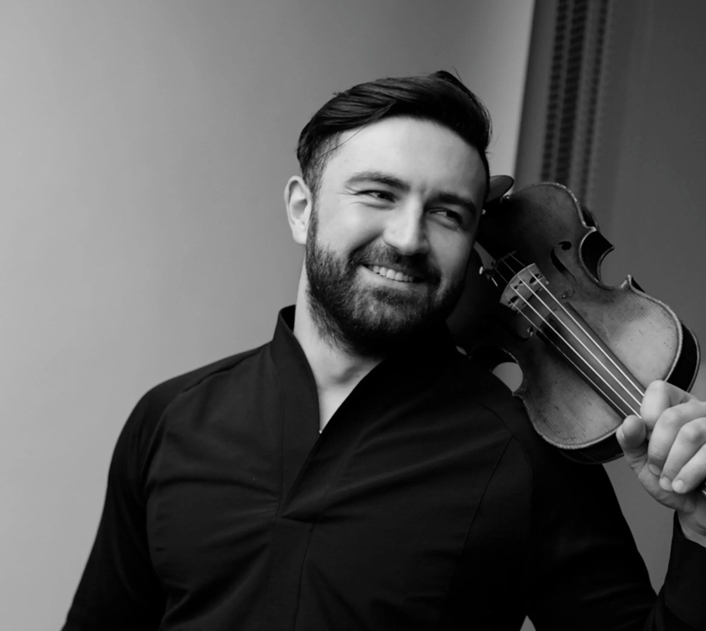 Black and white photo of a man with a beard smiling, holding a violin close to his face. The background is plain and light-colored.